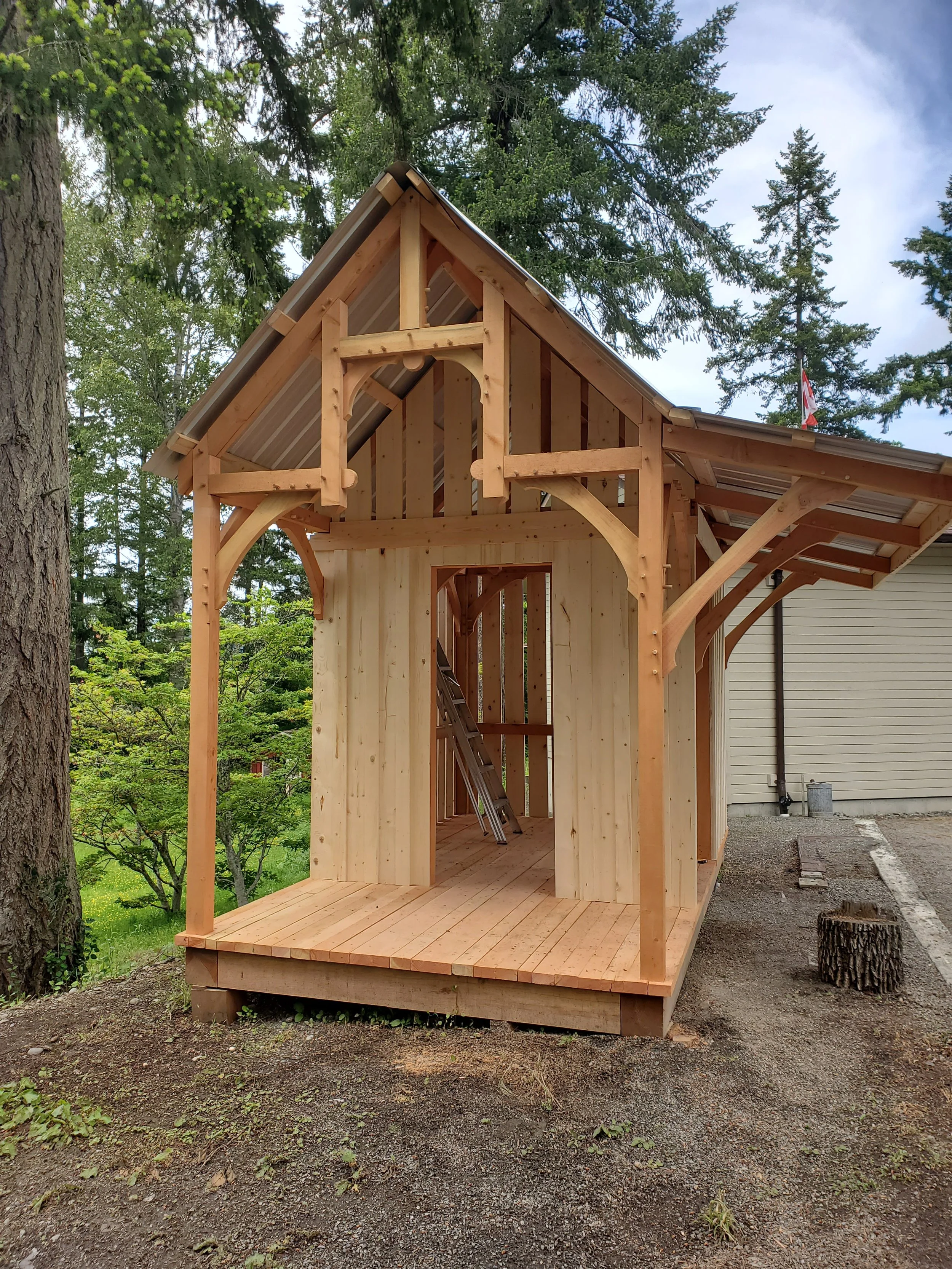 A wooden Bunkie shed under construction, with an open ground floor and a pitched roof, surrounded by trees and a cloudy sky.