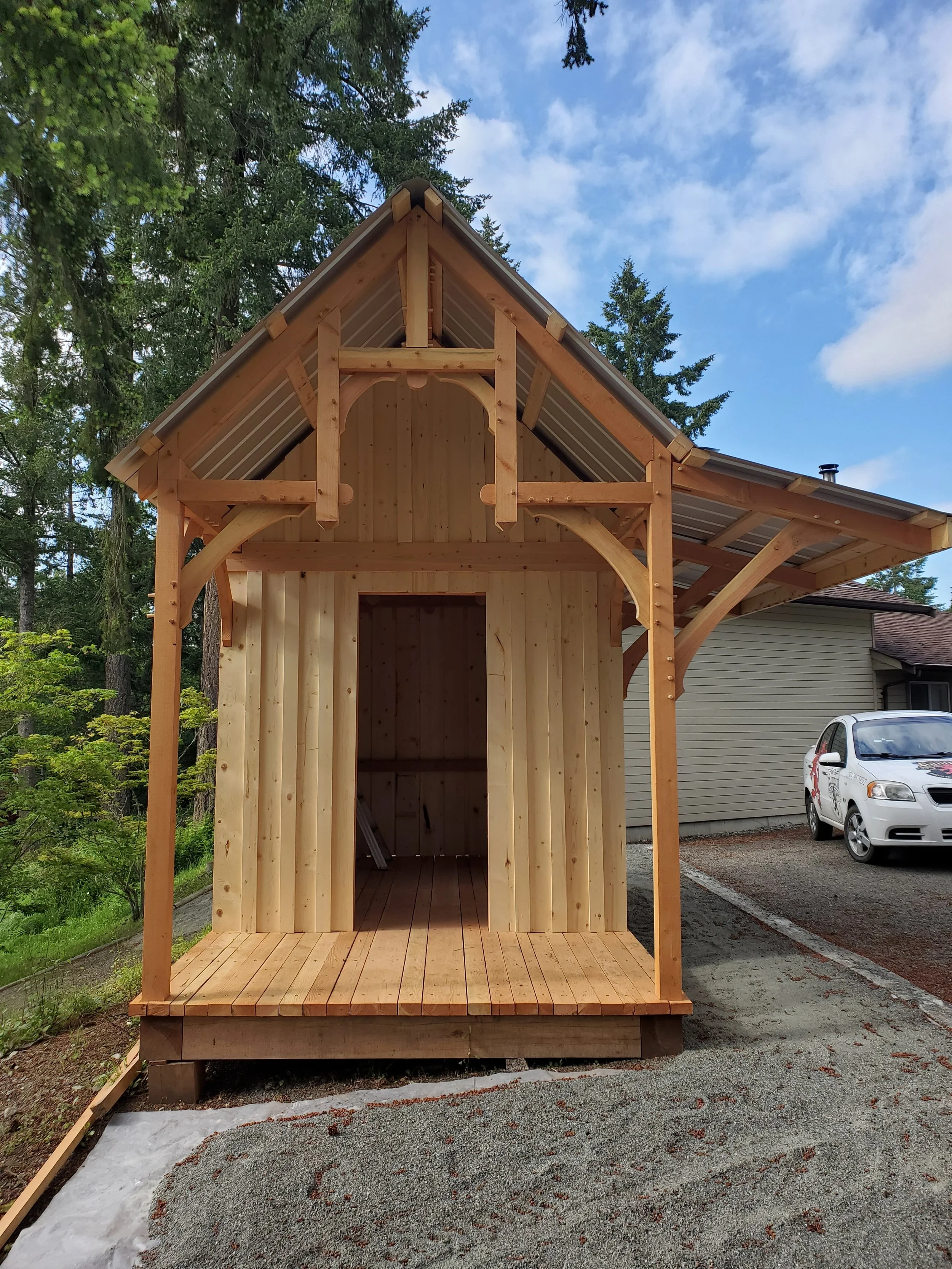 A wooden structure under construction with a partially finished upper section, set outdoors near trees and a house. There is a white car parked on the right side of the image.