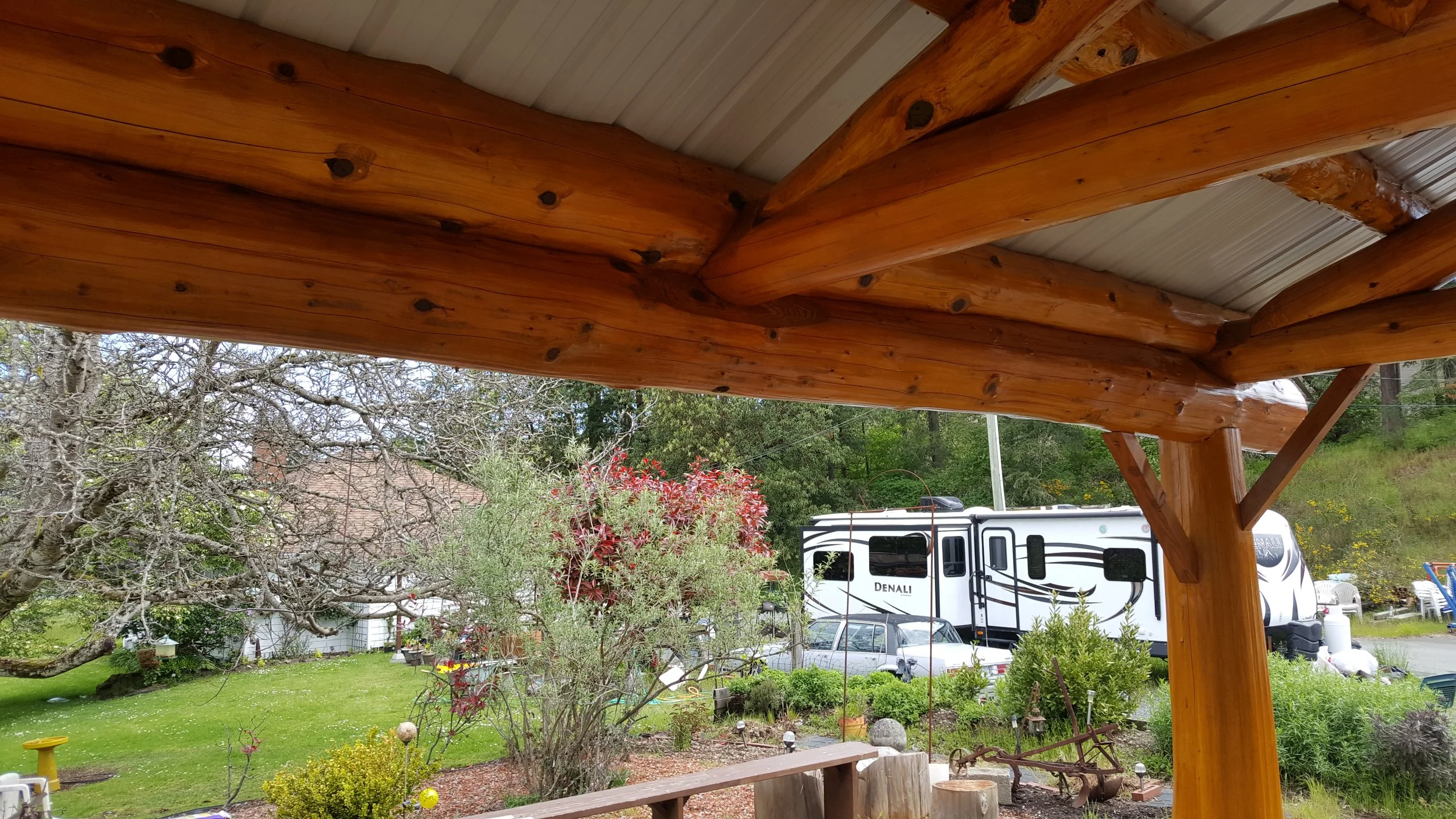 Wooden porch ceiling with large logs and metal roof panels, overlooking a lush garden with trees, bushes, and an RV parked in the background.