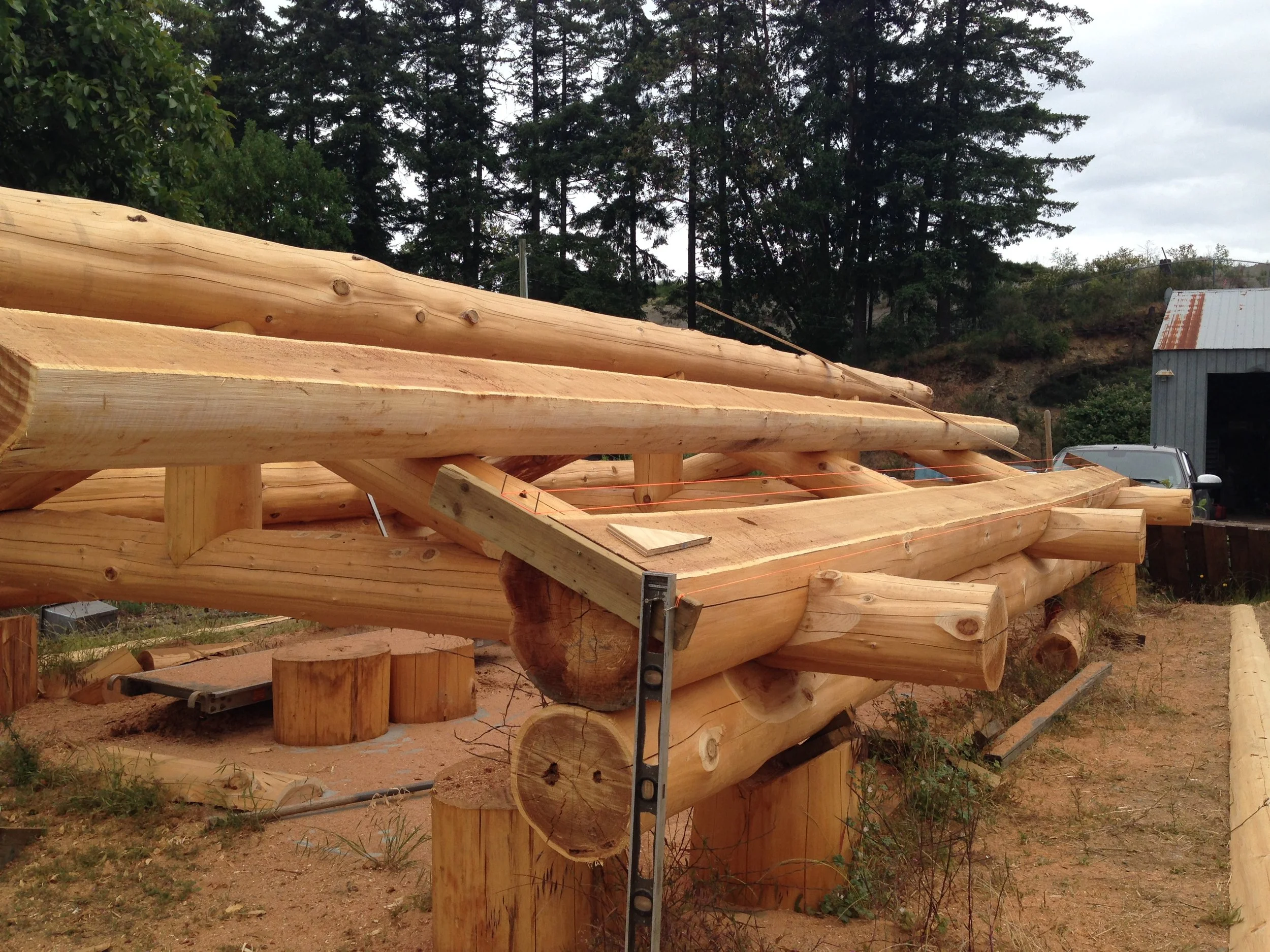 Wooden log structure under construction outdoors, with trees and a gray building in the background.