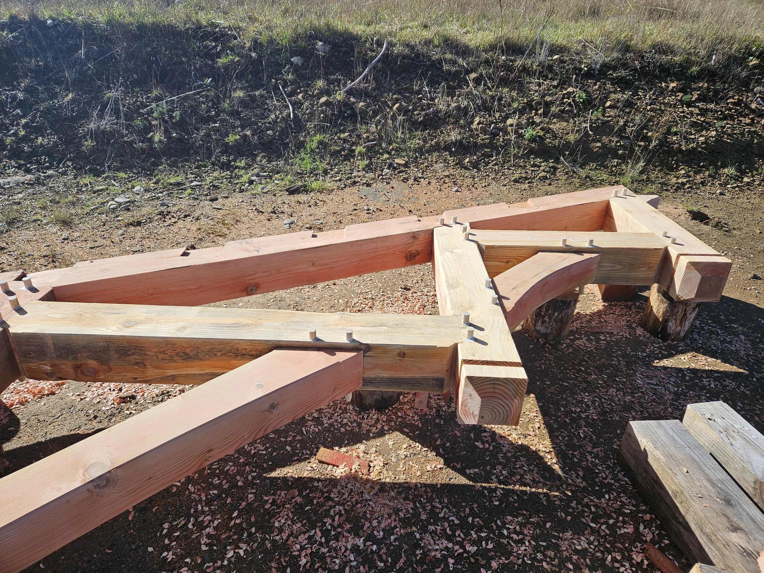 Close-up of a wooden structure being assembled outdoors on dirt and gravel ground, with grassy hillside in the background.