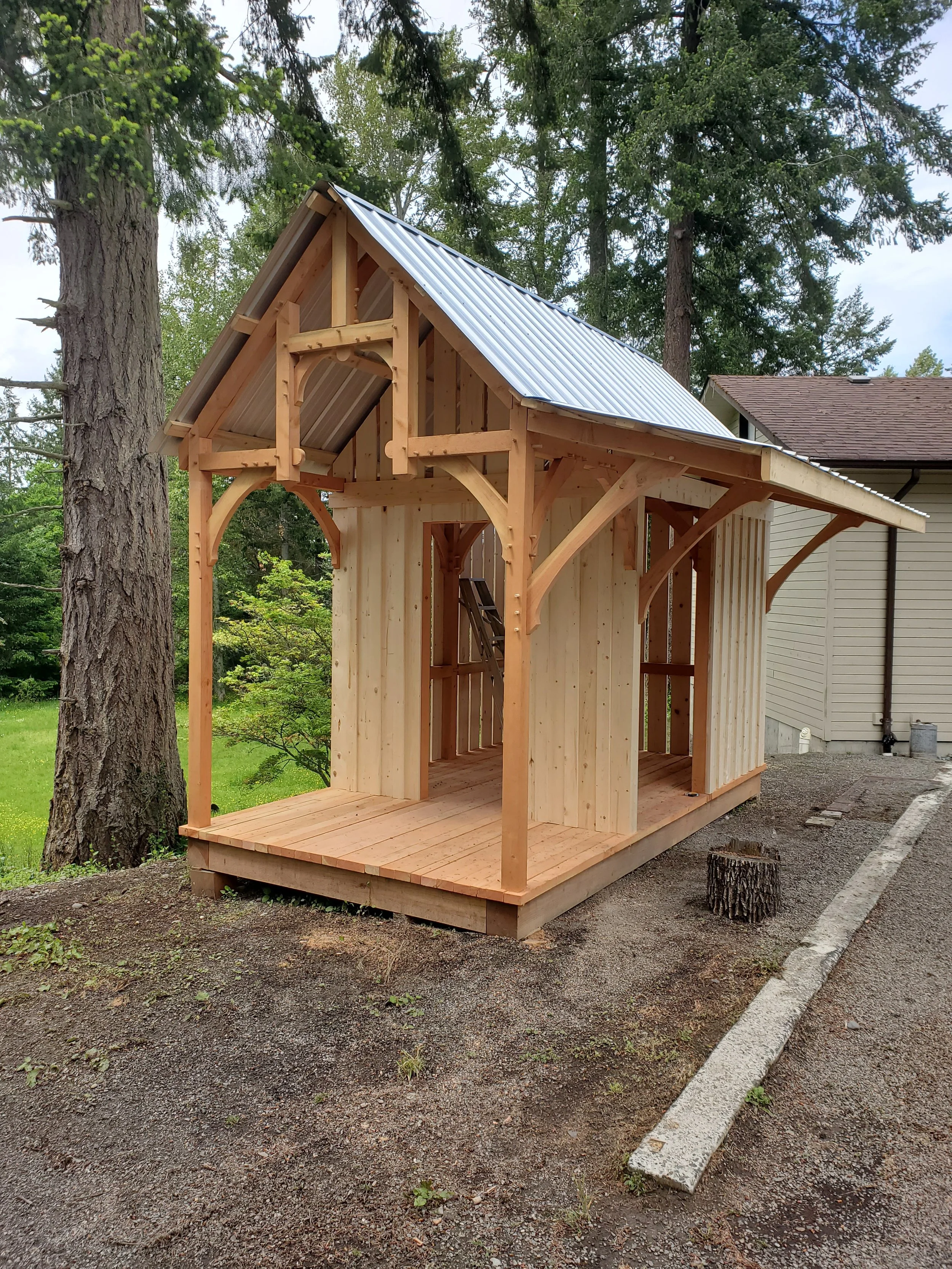 Unfinished wooden bunkie shed with a metal roof, surrounded by trees and a house in the background.