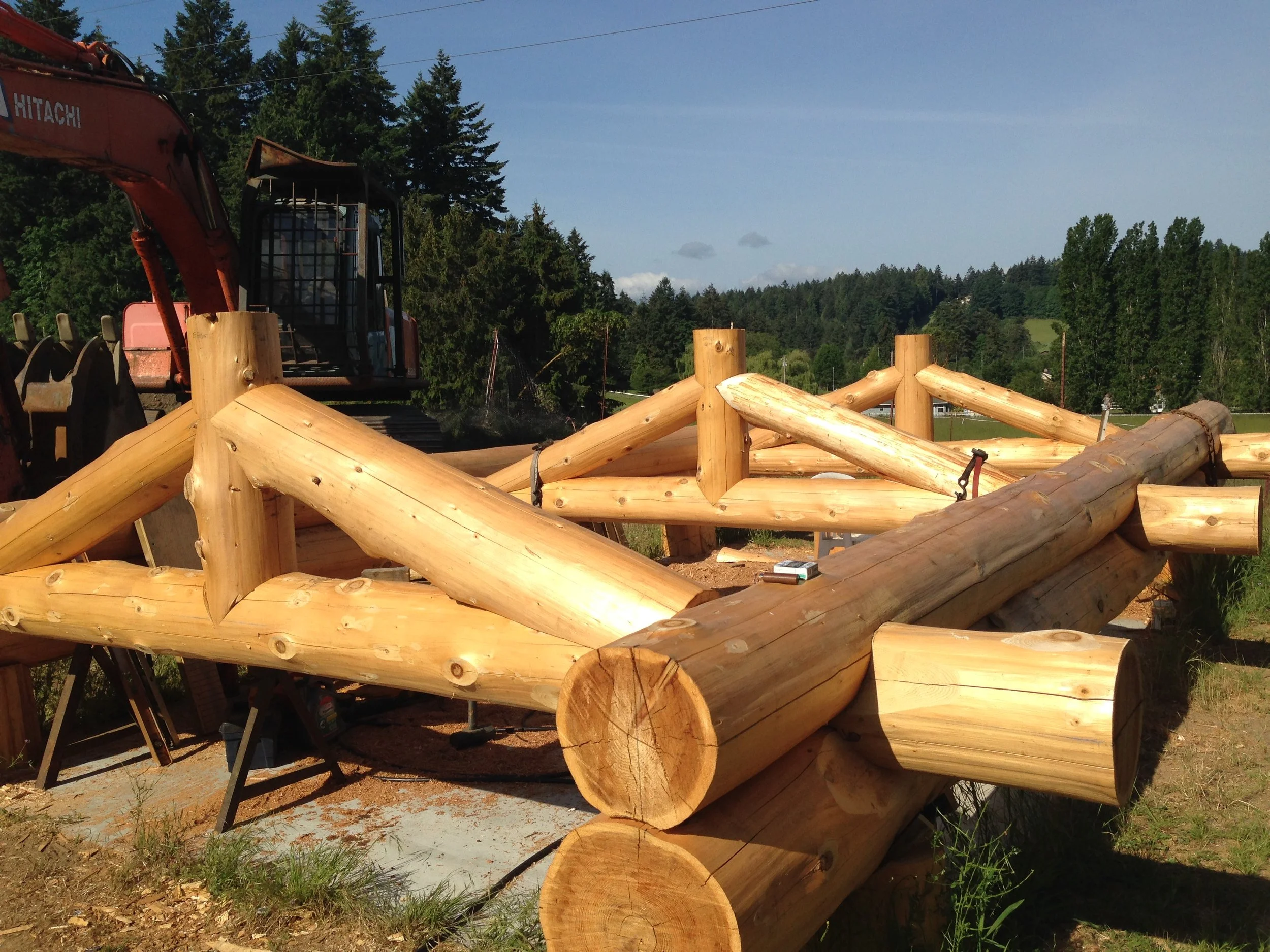 A construction site with large wooden logs arranged in a structure and a red Hitachi excavator in the background, surrounded by trees and a blue sky.