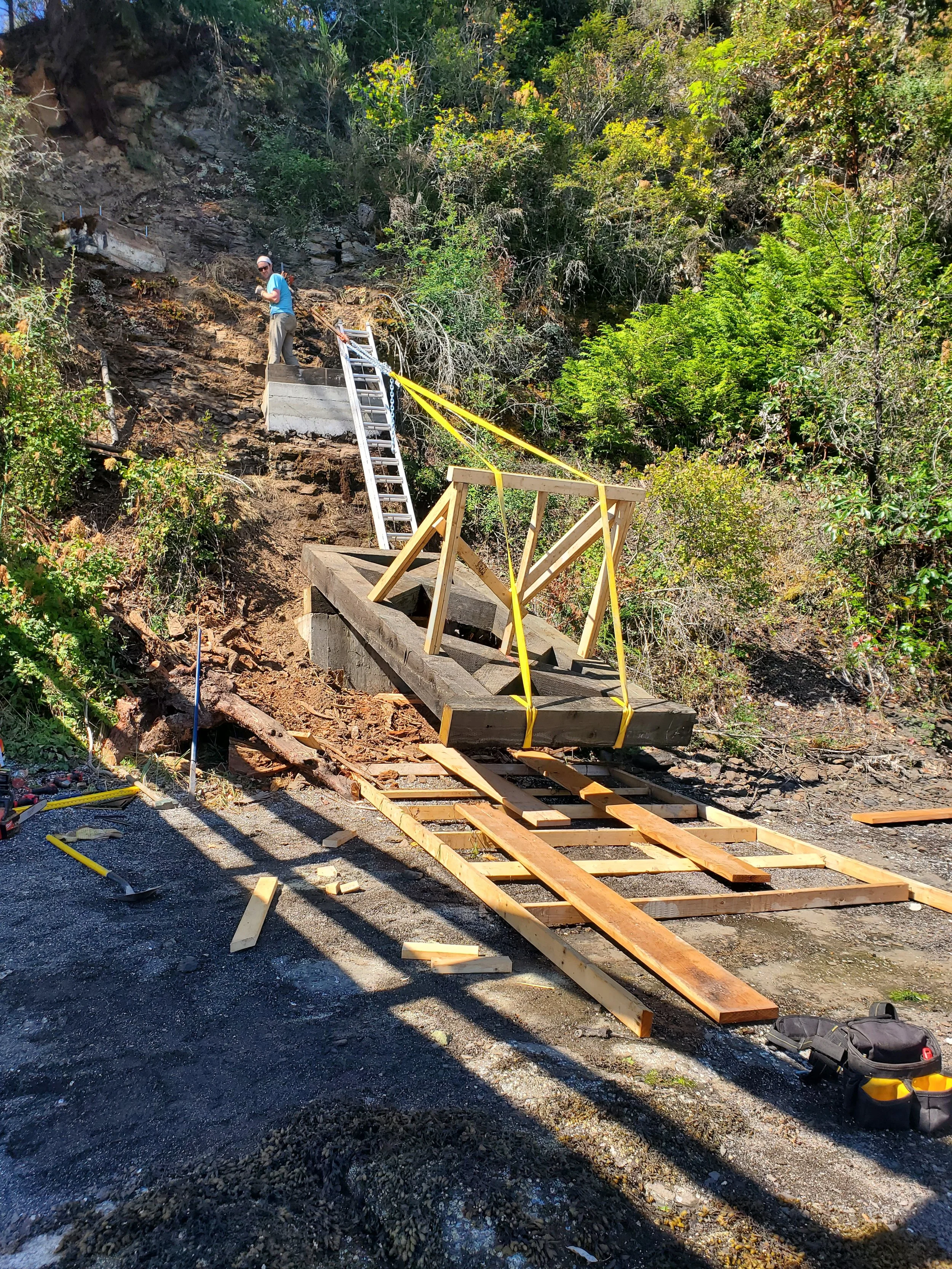 A person standing on a construction site on a hillside with wooden framing and scaffolding for building a bridge or similar structure, surrounded by trees and outdoor environment.