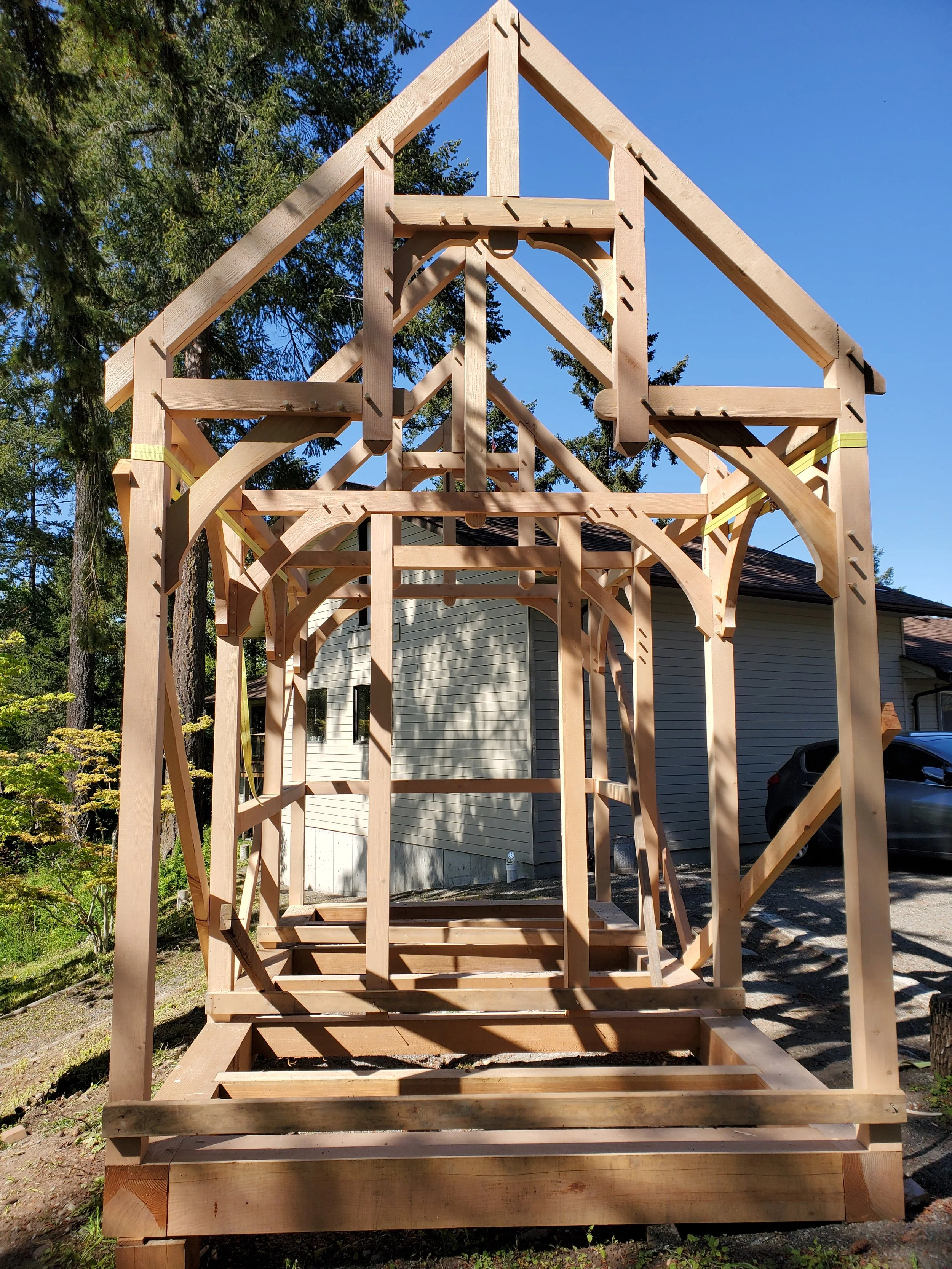Wooden framing of a house under construction, with a clear blue sky and some trees in the background.
