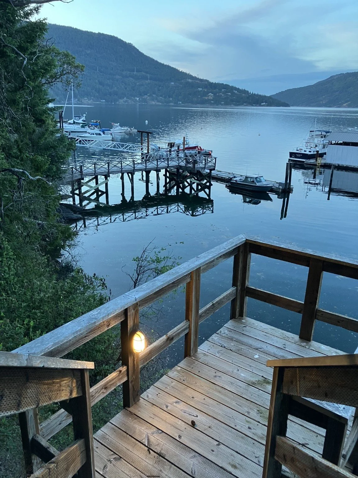 A wooden deck, and stairs overlooking a calm lake with boats docked at a pier and lush green mountains in the background during dusk.