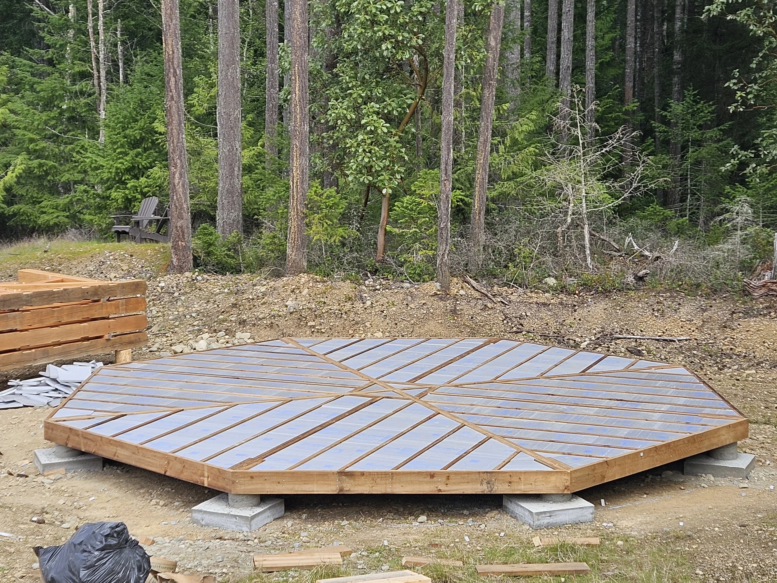 Construction site with a wooden platform raised on concrete blocks, surrounded by gravel and dirt, with trees and a forested area in the background.