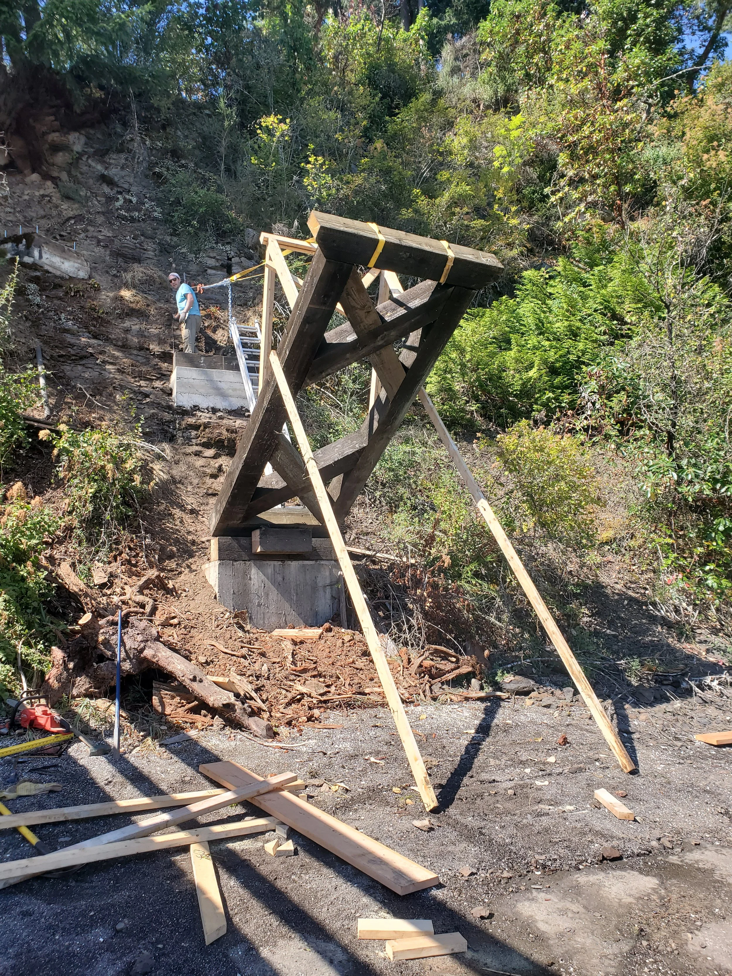 A construction site on a hillside, with a person working among wooden structures and tools, surrounded by trees and greenery.