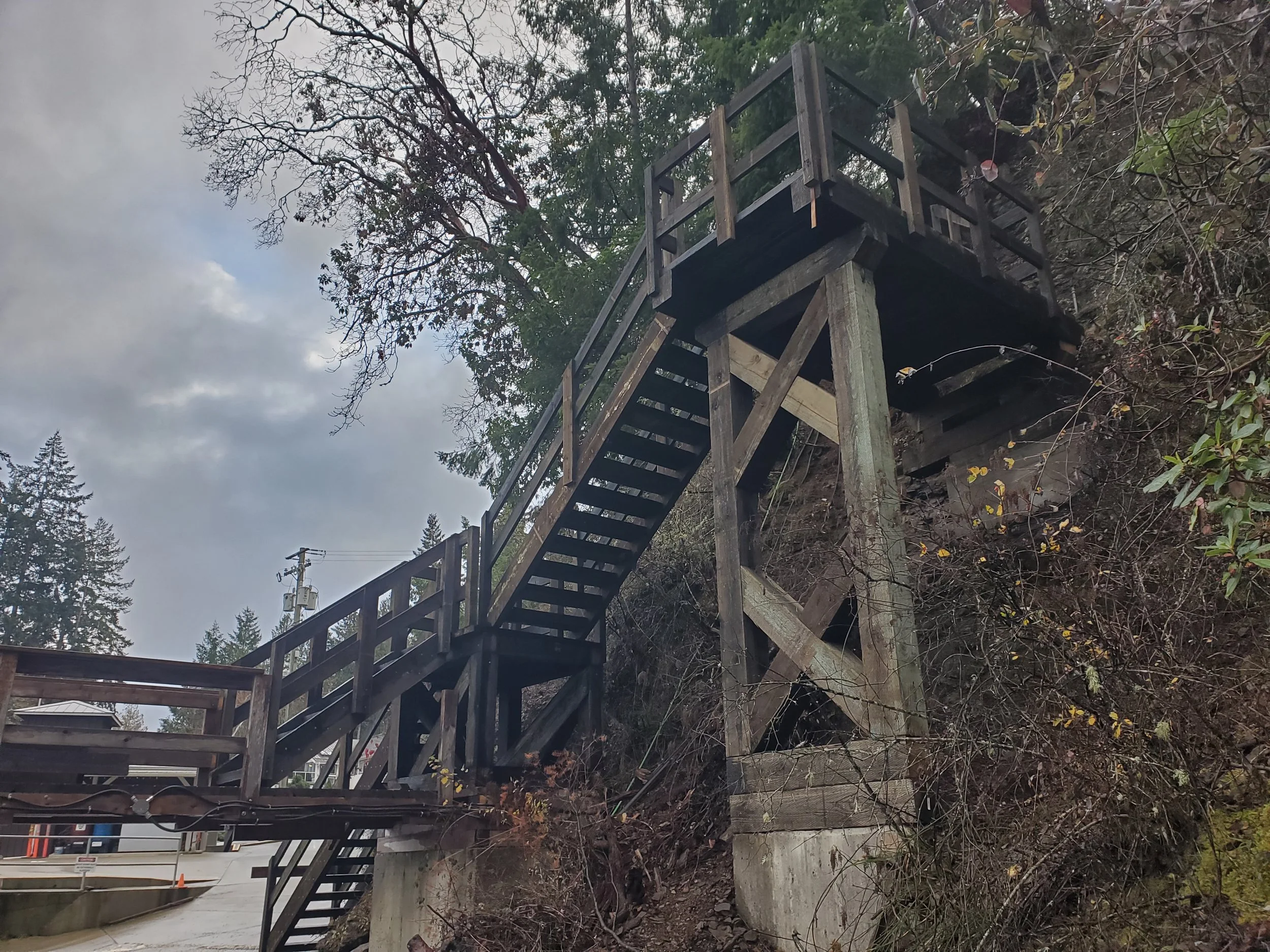 A wooden staircase with railings rising alongside a slope covered in bushes and trees. The sky is overcast with some clouds. In the background, utility poles and a road are visible.