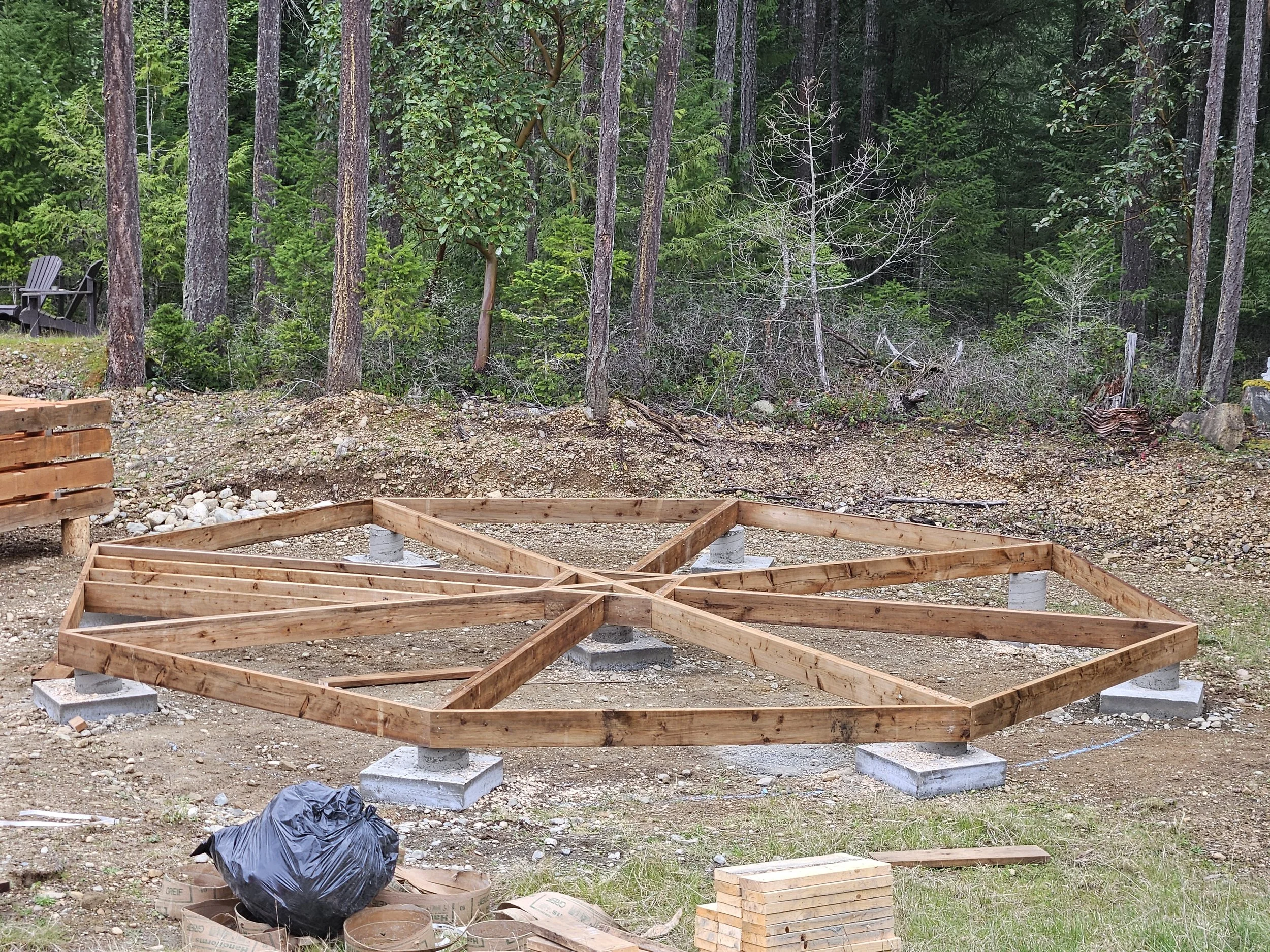Starting framework for a small structure, with circular wooden outlines on concrete footings, surrounded by construction tools and materials. In the background, a dense forest of tall, green pine and other trees.