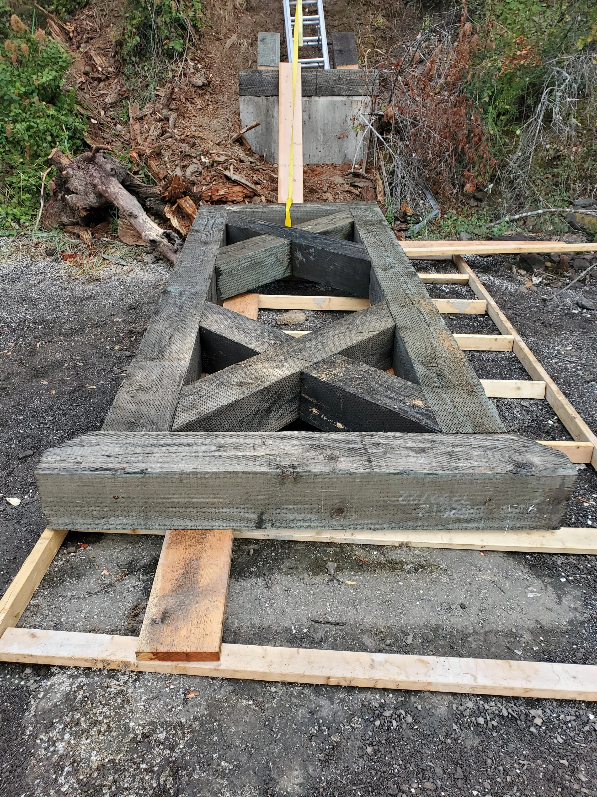 Construction site with wooden framing for a foundation or deck being built, with some blackened wood and a ladder at the far end, surrounded by dirt and vegetation.