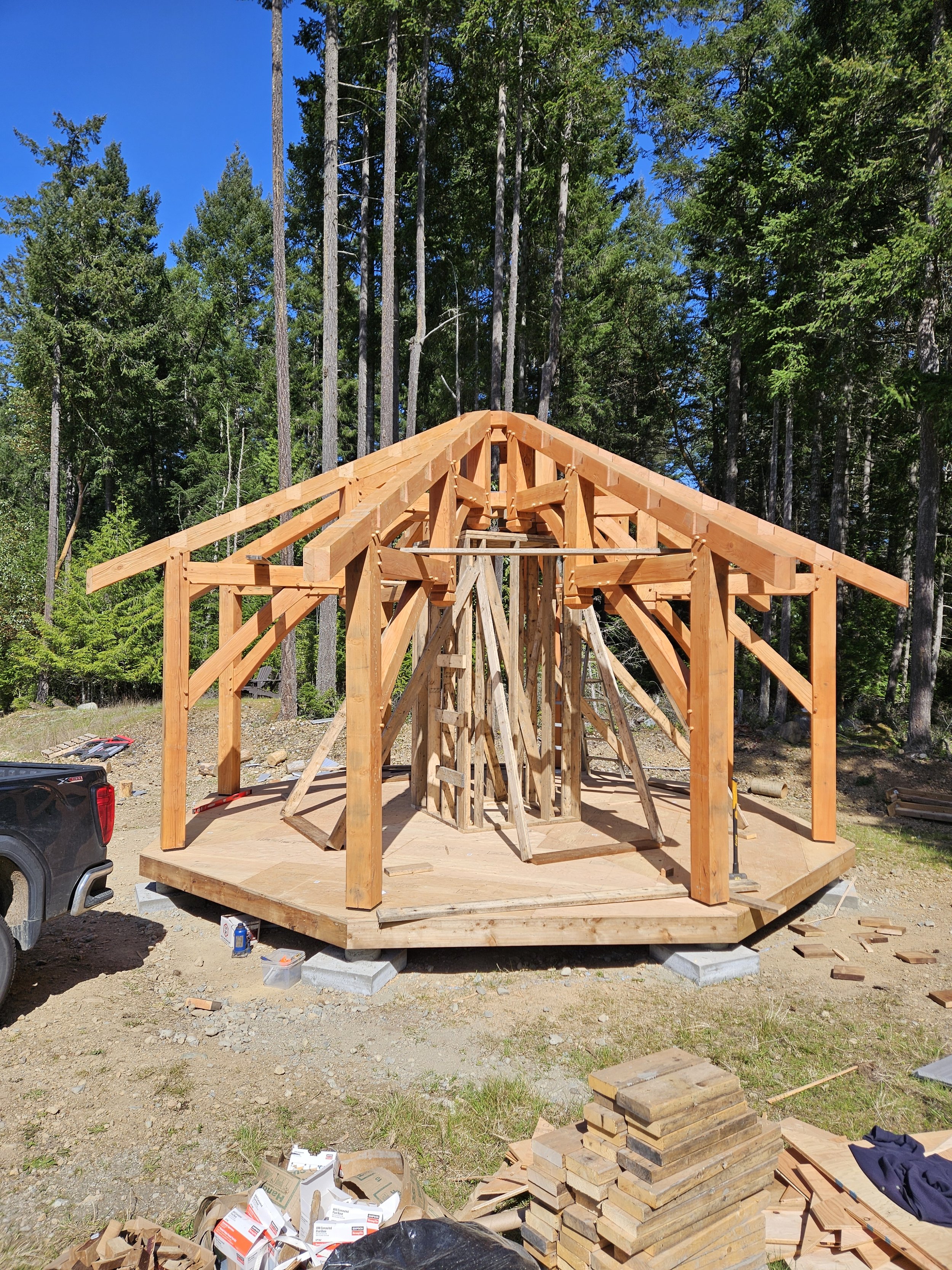 Wooden structure under construction in a forested area with trees and blue sky in the background, construction tools and materials scattered on the ground.