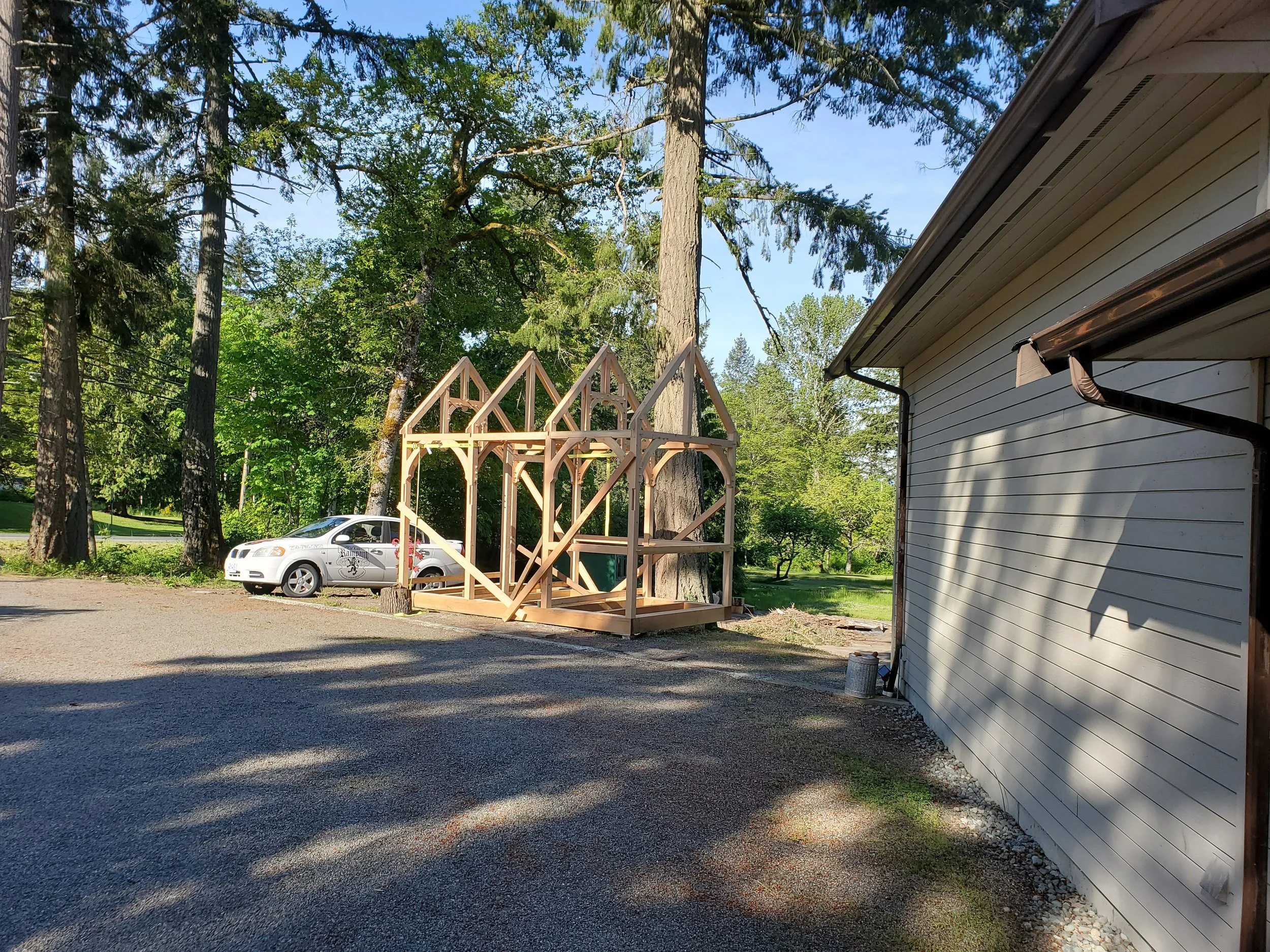 Wooden framing for a house construction in progress next to a white house, with tall trees and a parked car in the background.