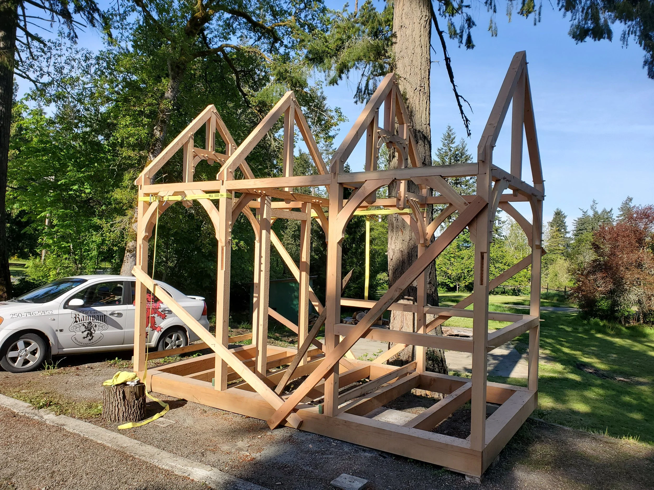 Wooden house frame under construction next to a tree, with a car parked nearby and lush green trees in the background.