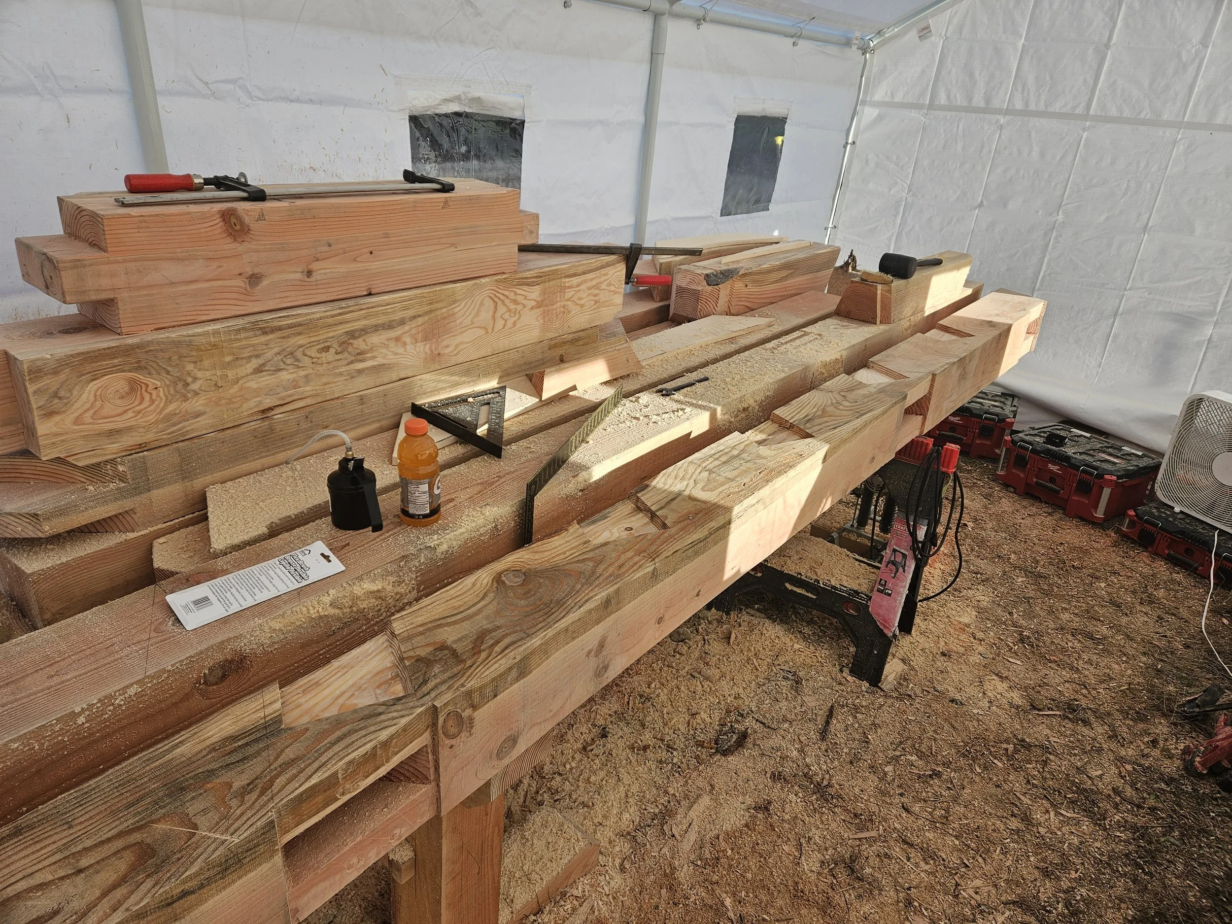 Construction site with wooden beams arranged for a project inside a tent. Various tools, including clamps and a mallet, are placed on the beams. The ground is dirt, and there are some power tools and a small fan in the background.