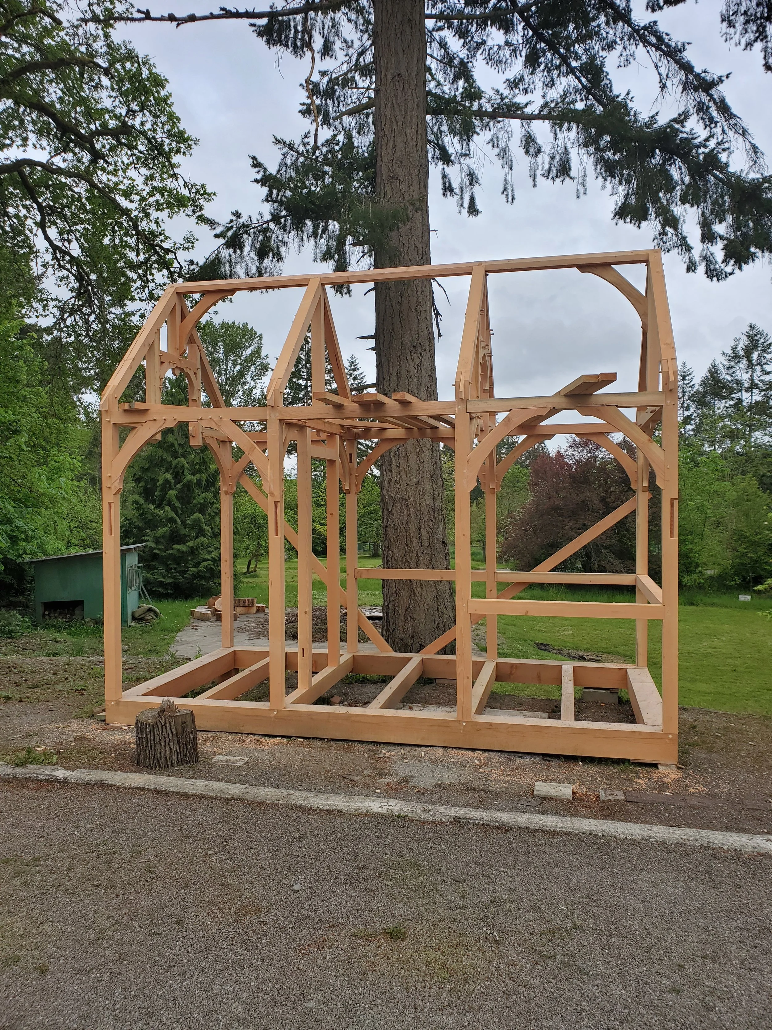 Wooden framing structure under construction outdoors with trees and cloudy sky in the background.