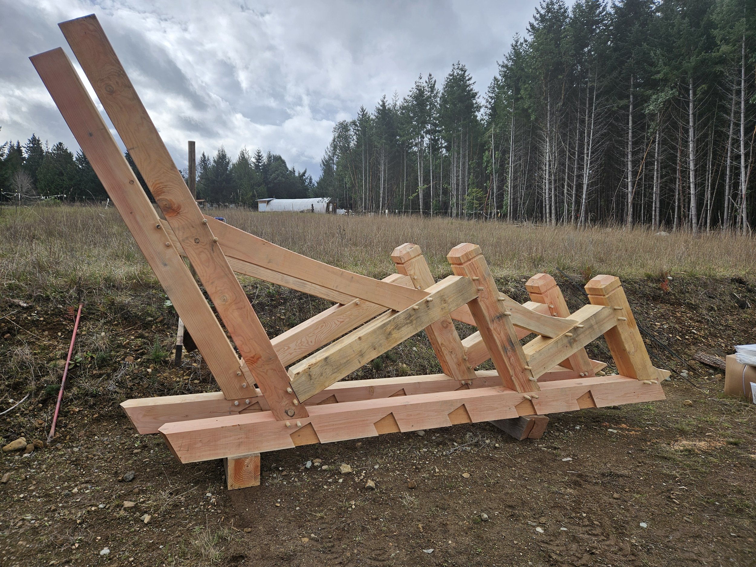 Wooden framework construction site in a field with a forest in the background and cloudy sky.