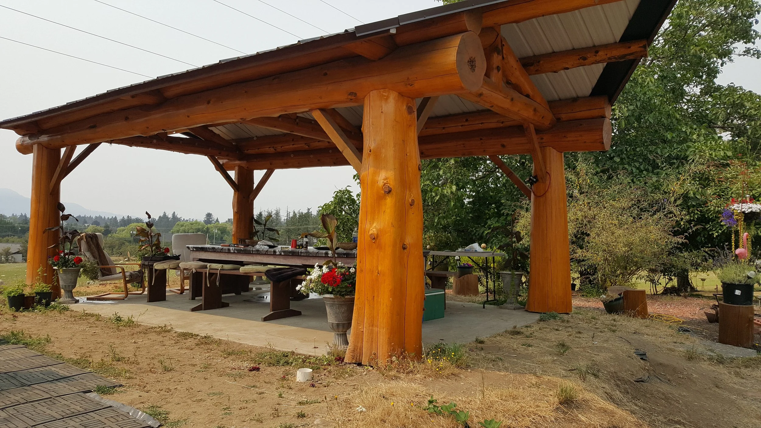 A wooden pergola with a metal roof, surrounding a patio with outdoor seating, potted plants, and a table, in a garden setting with trees and landscape in the background.