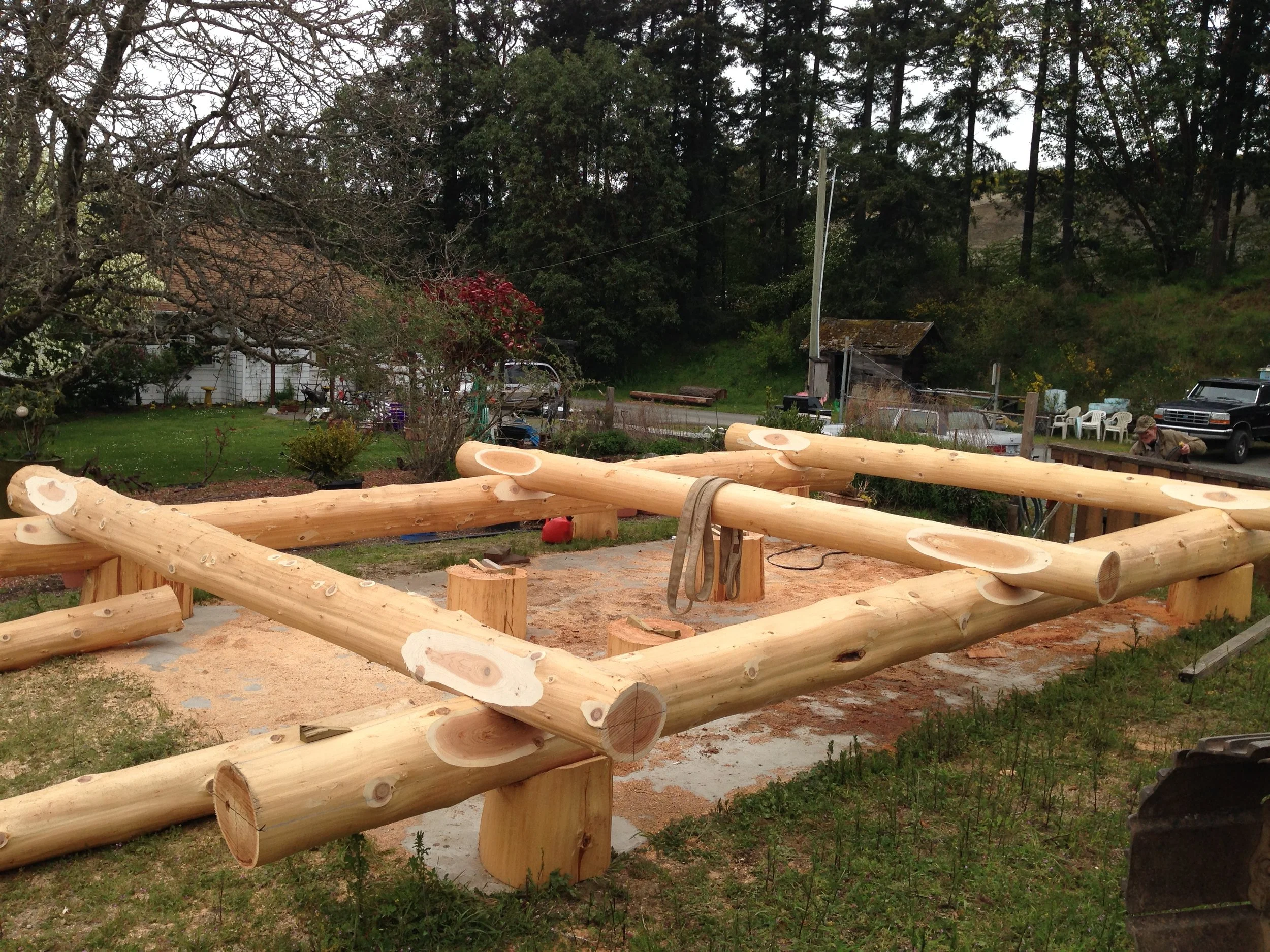 A woodworking area with large logs and a partially constructed wooden structure in the foreground, with a yard and trees in the background.
