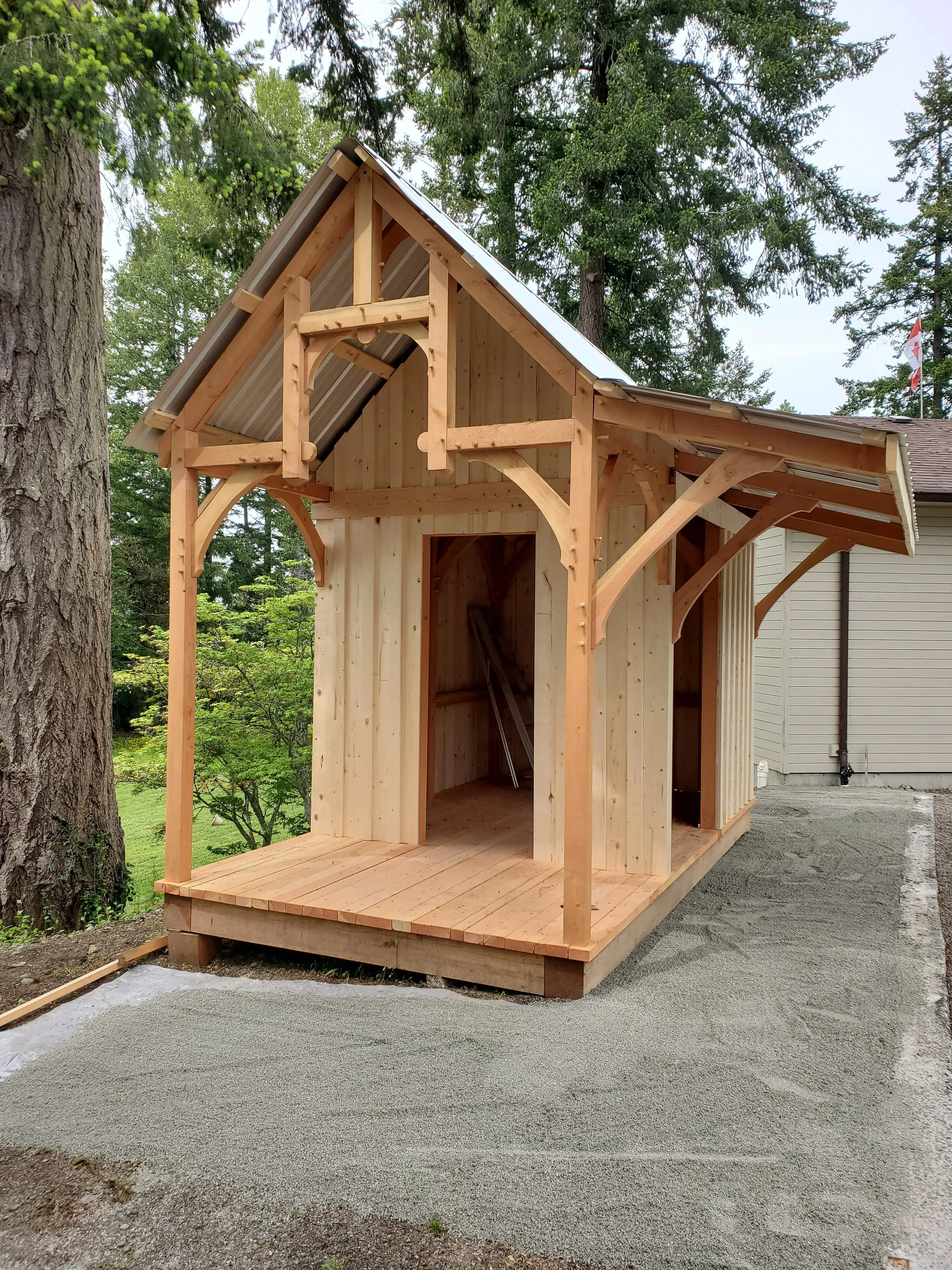 A partially constructed wooden shed or small building with a pitched metal roof, situated outdoors among trees and next to a house with siding, with gravel on the ground in front.