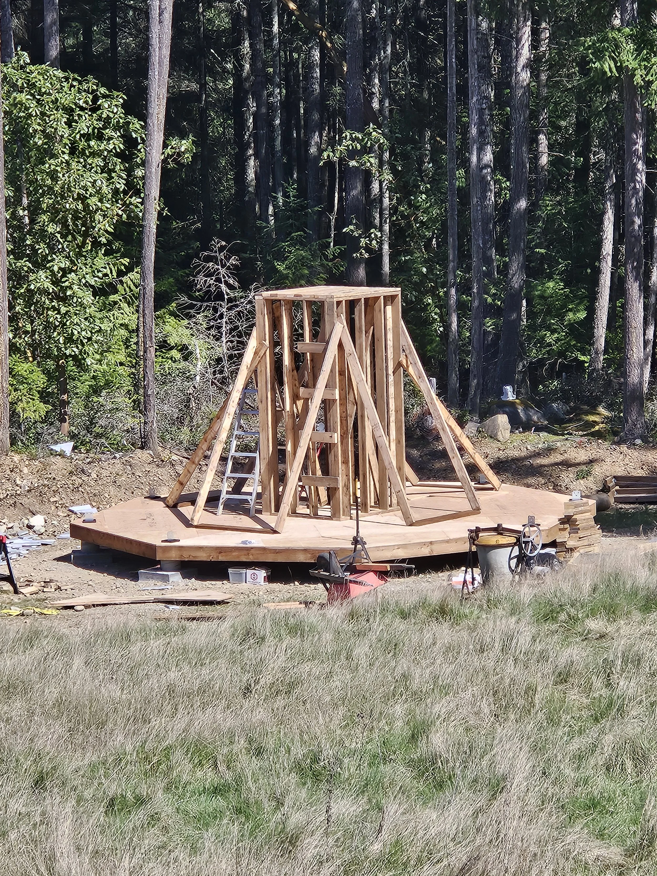 A wooden construction frame is being built on a circular wooden platform in a forest clearing, surrounded by trees and grass.