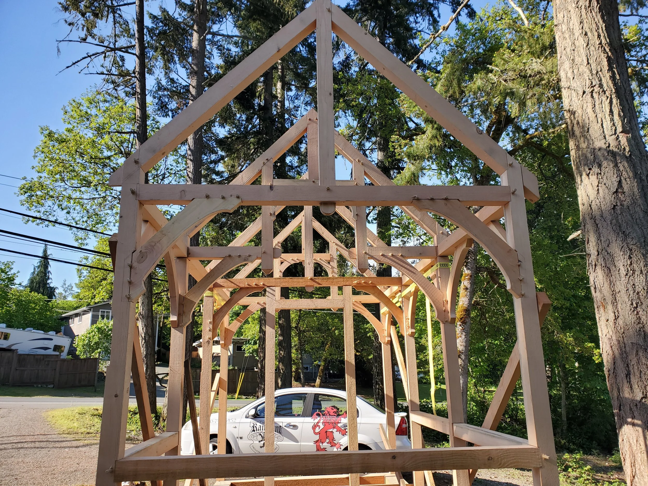 Wooden framing structure for a house or building under construction in a backyard with trees, a car, and a house in the background.