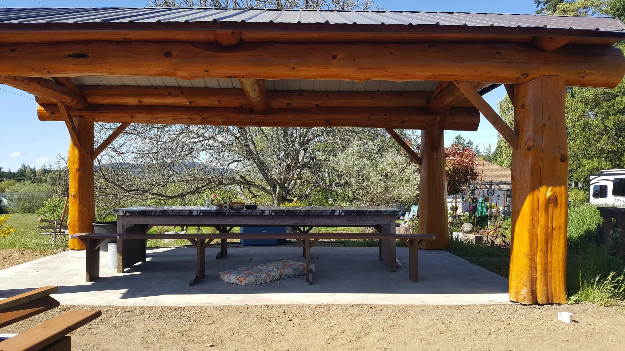 A wooden outdoor shelter with a metal roof, featuring a picnic table underneath, surrounded by trees and greenery, with a clear blue sky in the background.