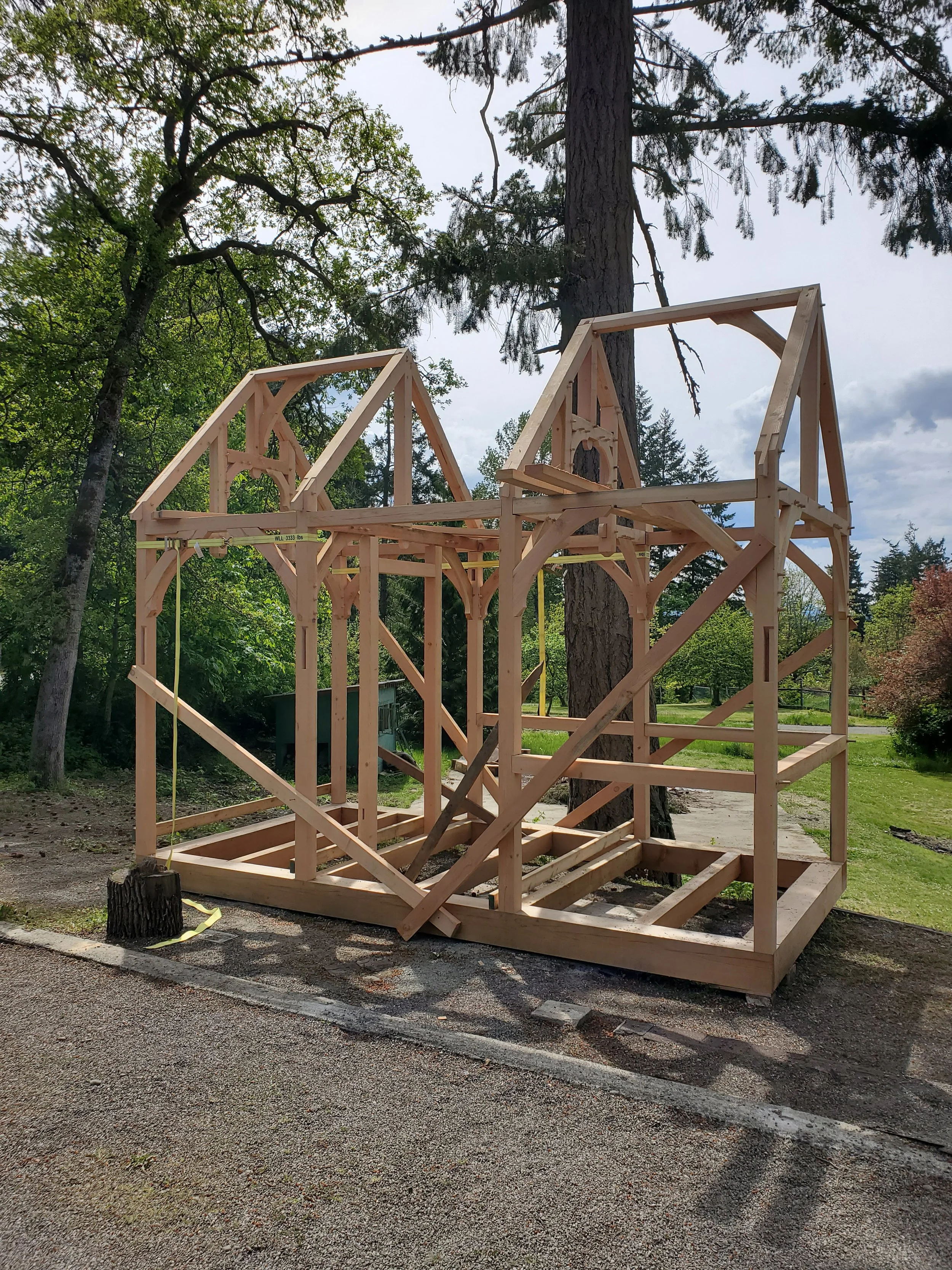 Wooden frame structure under construction in a park, surrounded by trees and greenery, with a partly cloudy sky.