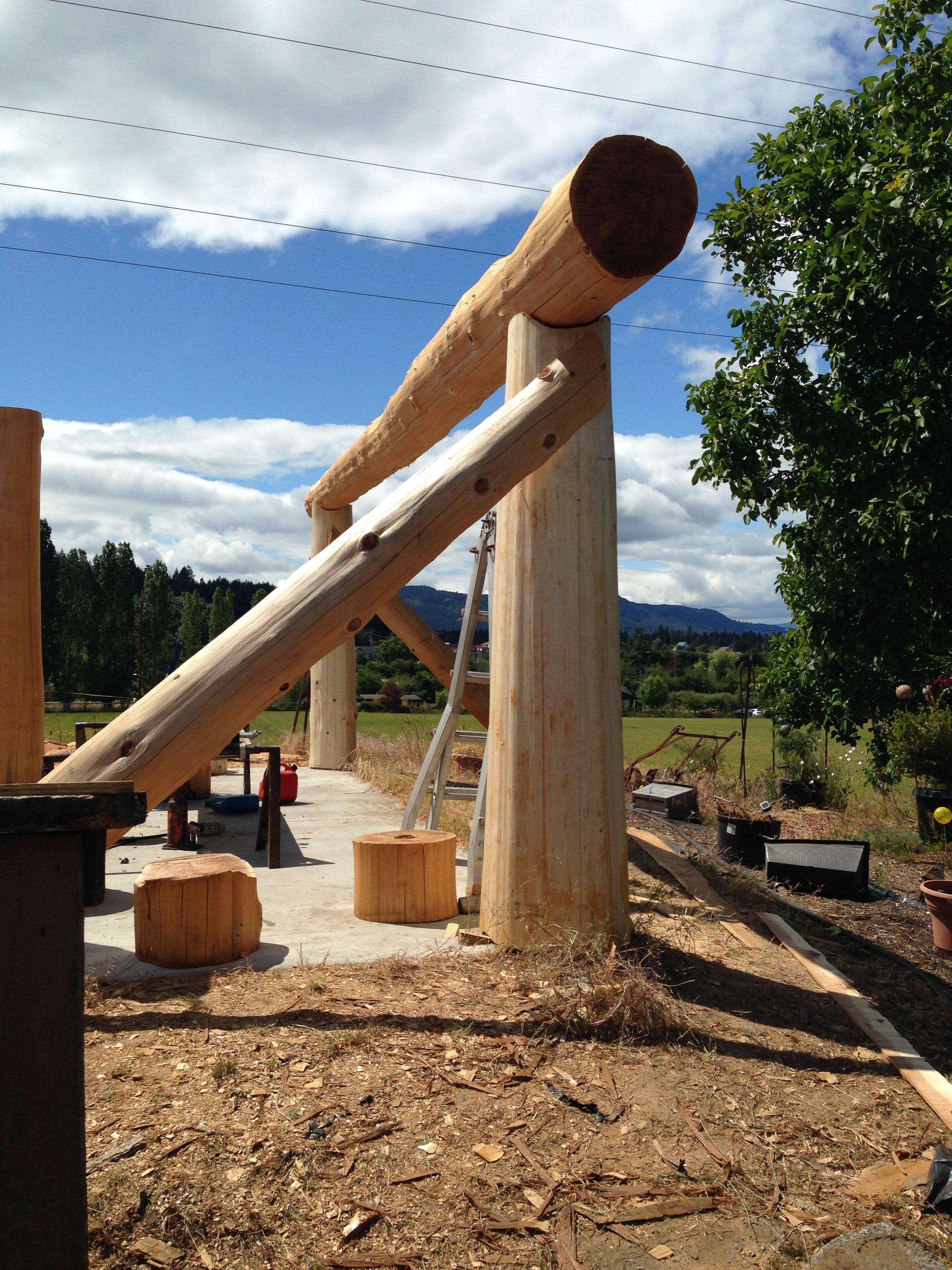 Wooden logs are being assembled to construct a large outdoor structure, with a lush green tree and cloudy sky in the background.