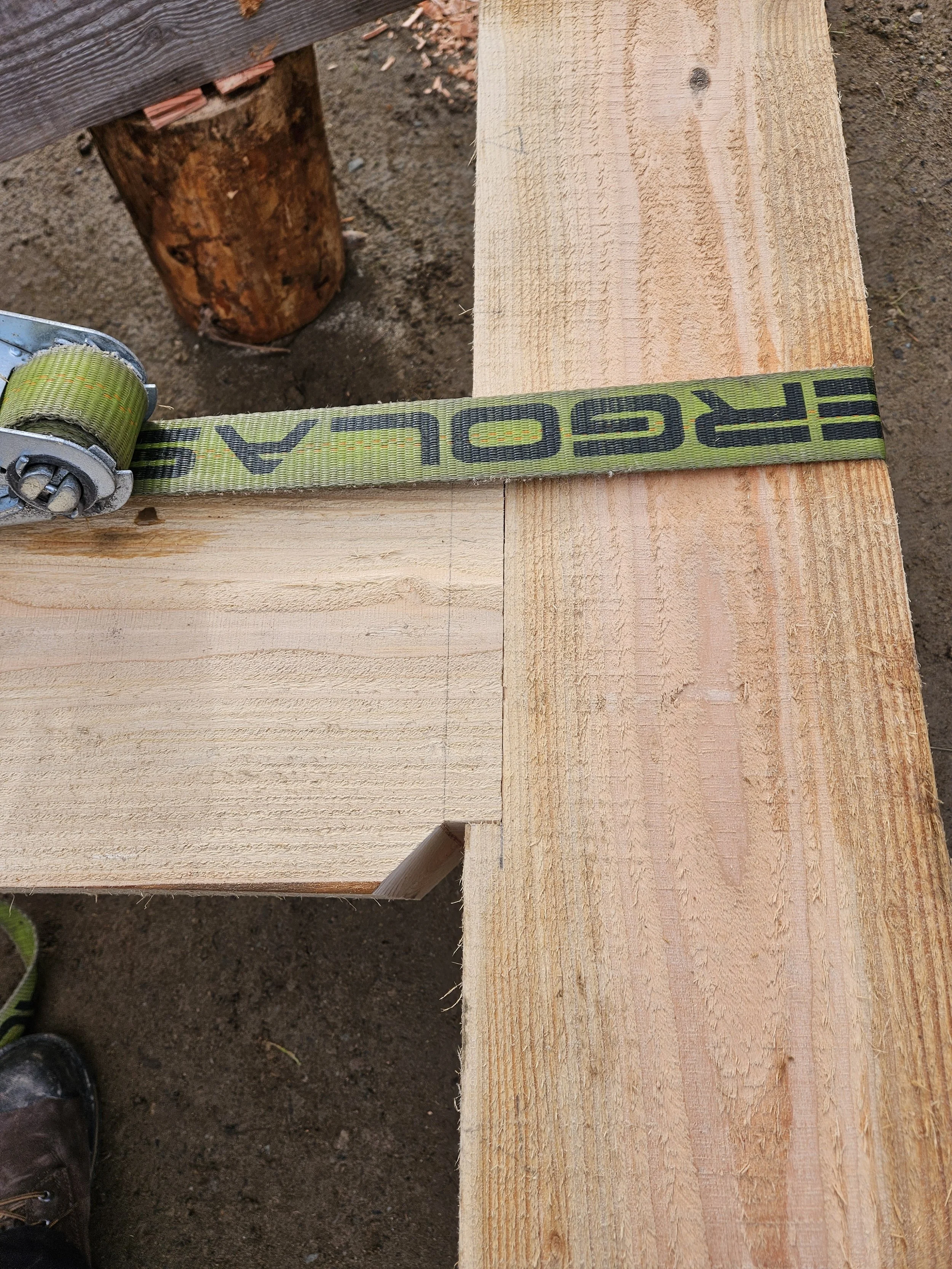 Close-up of wooden planks and a ratchet strap in a construction setting, with dirt and a person's shoes visible at the bottom.