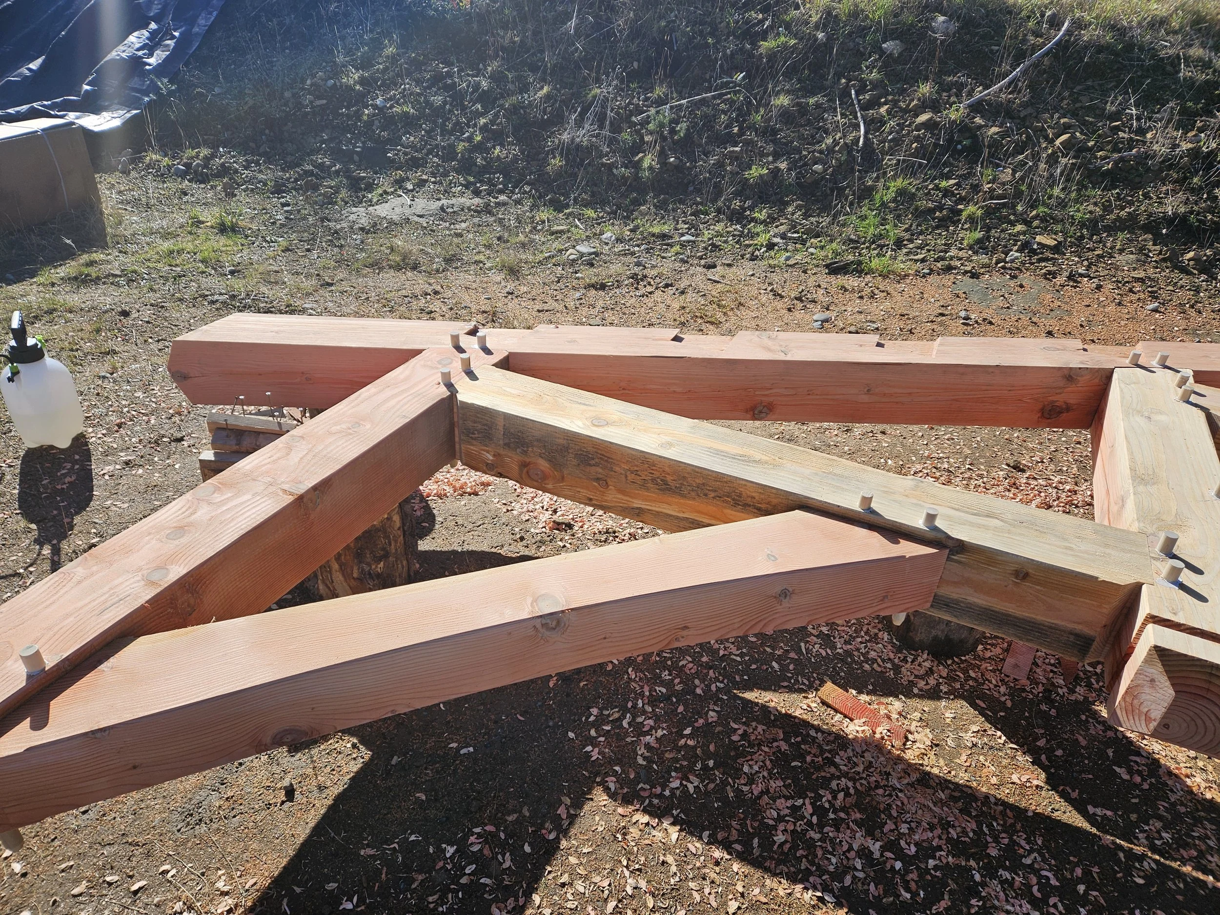 Close-up of a wooden structure under construction with bolts, on uneven ground with small rocks, dirt, sparse green plants, and background of rocky soil and dried grass.