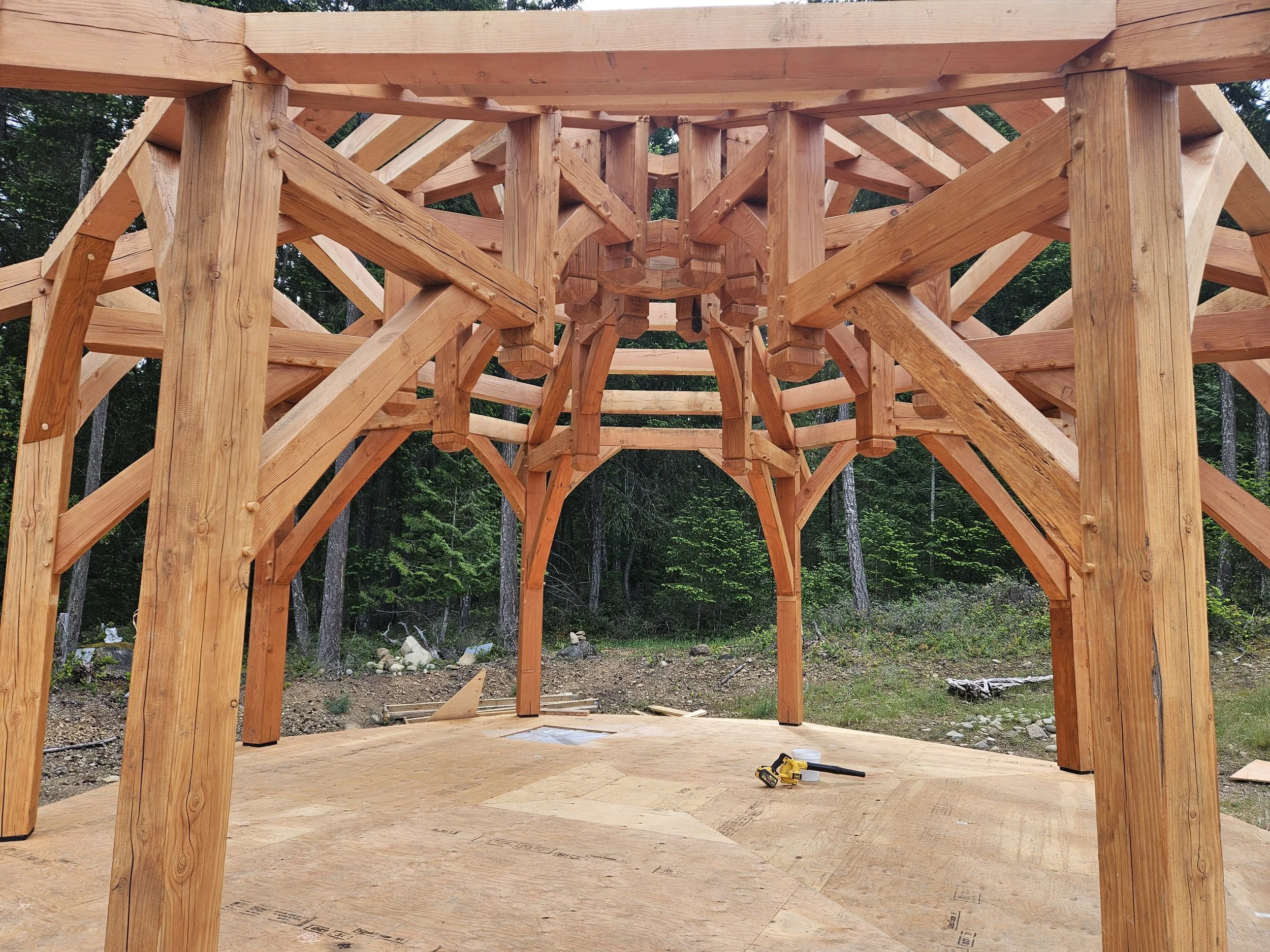 The photo shows the interior of a house under construction, with a wooden framework and a plywood floor, in a forested area.