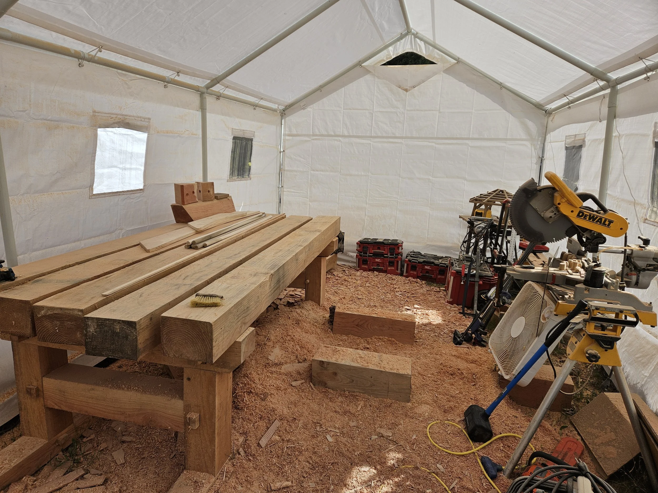 Interior of a construction site with wooden workbench, building materials, and power tools including a circular saw inside a temporary shelter with white fabric walls and ceiling.