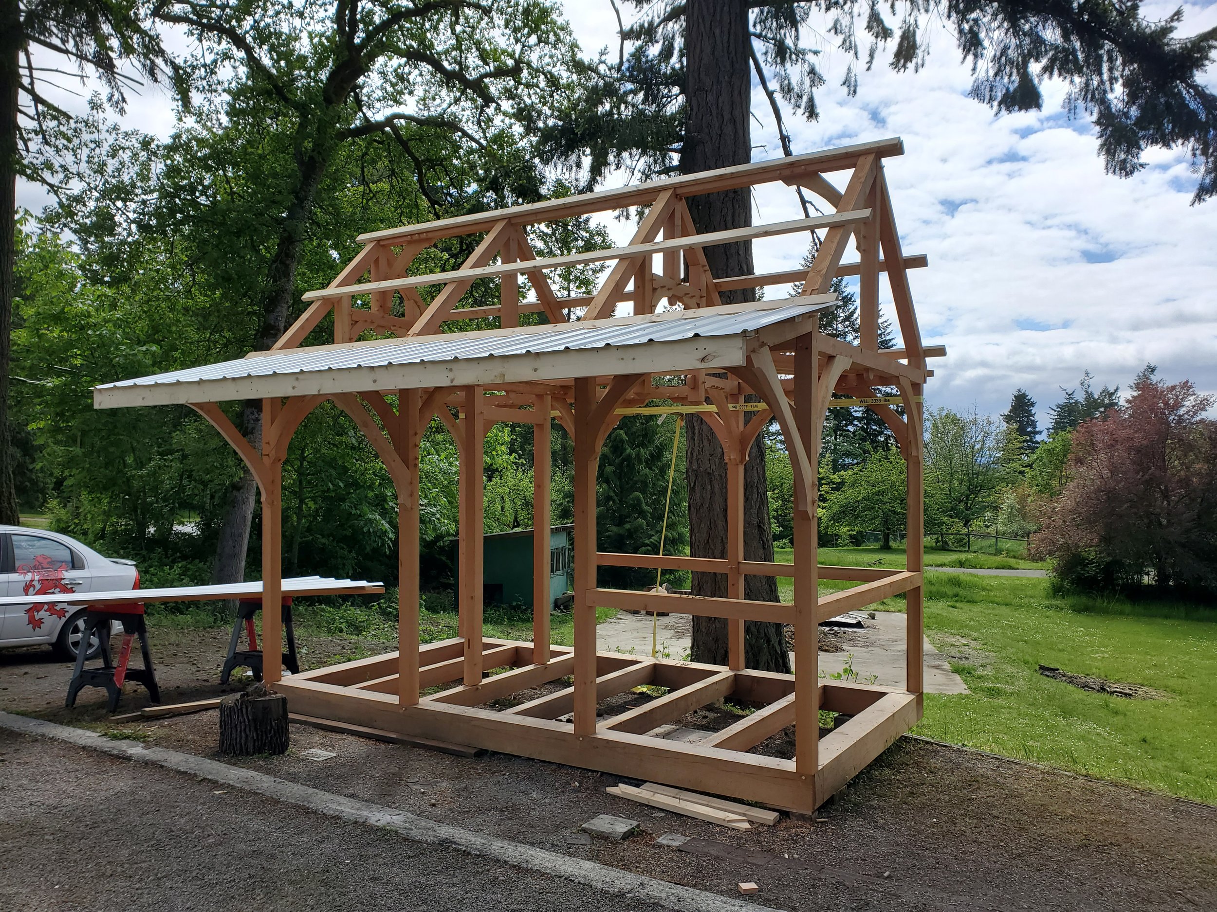A wooden framework for a timber bunkie is under construction in a forested area with green trees and a partly cloudy sky in the background.