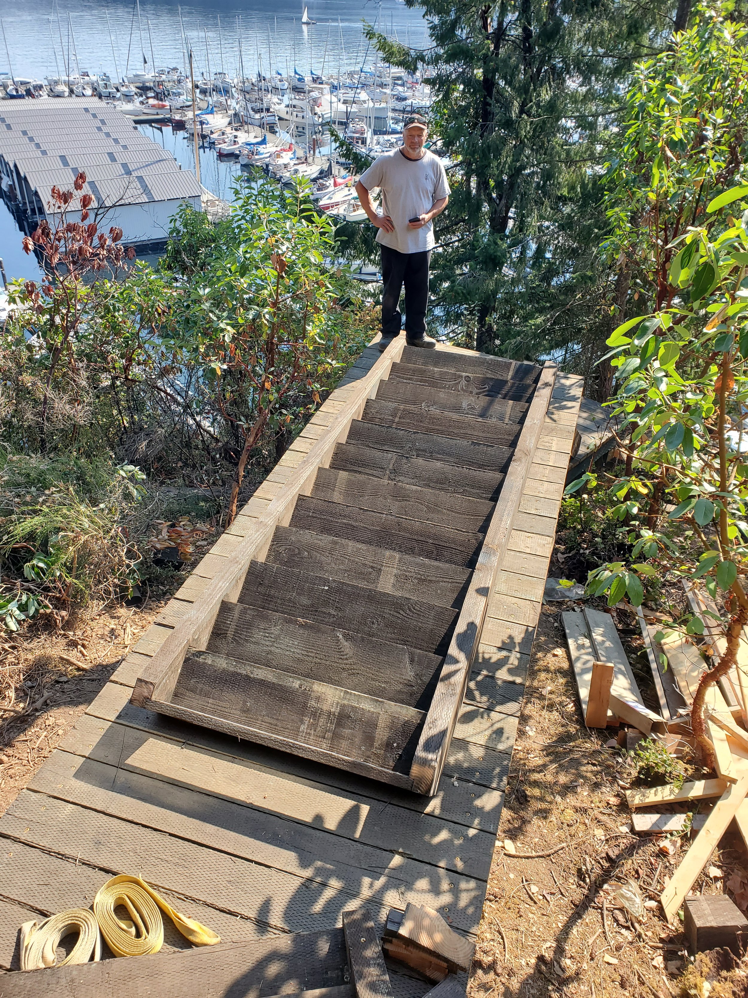 A man standing on a wooden staircase under construction near a marina with boats docked in the water.