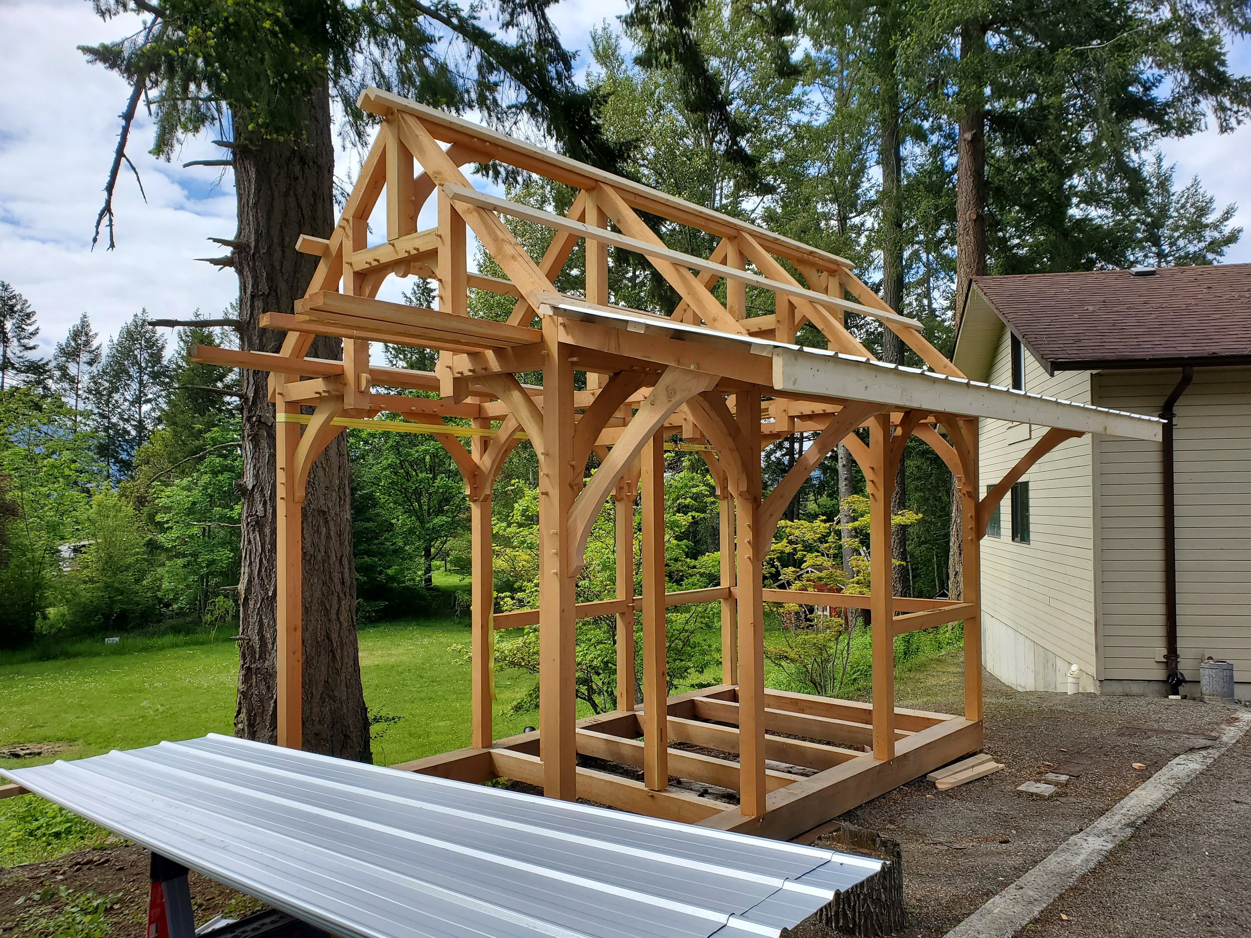 Wooden Bunkie frame under construction next to a house with a beige siding and brown roof, with trees and cloudy sky in the background.