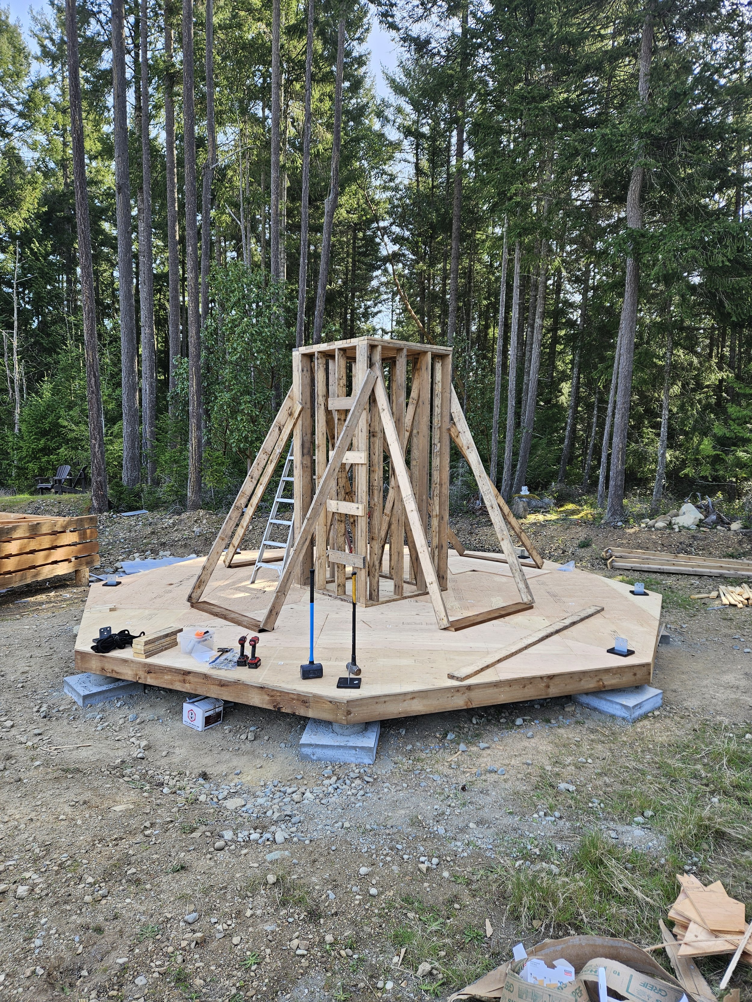Construction site in a wooded area where a wooden deck is being built. The structure’s foundation is supported by concrete blocks, and framing is in progress with wooden beams. Various tools and materials are scattered around the site, including scre