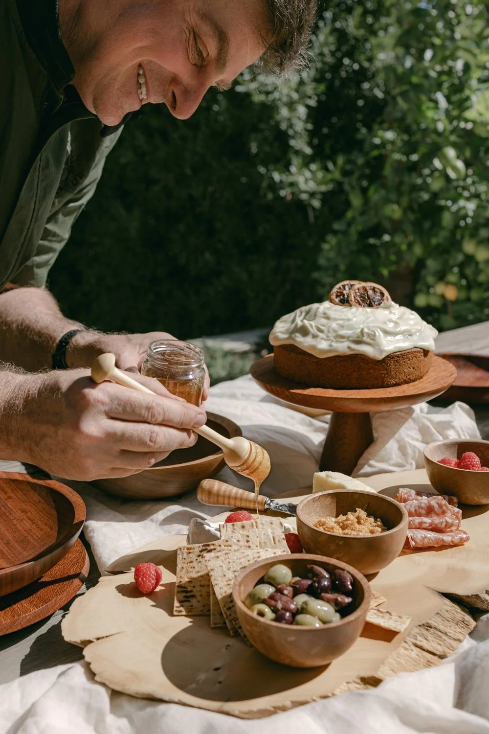 Andrew McNamara using a timber honey dipper over crackers on a charcuterie board surrounded by a table full of cheese, crackers, berries, and a cake topped with whipped cream and figs.