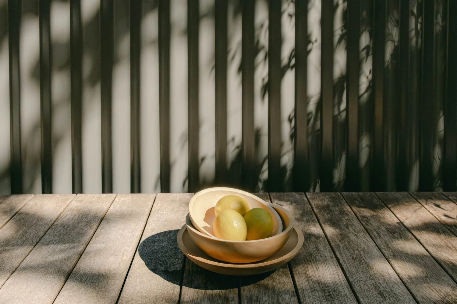 Two stacked timber bowls, the top bowl containing four green and yellow lemons, with shadows cast on the table and background by sunlight.