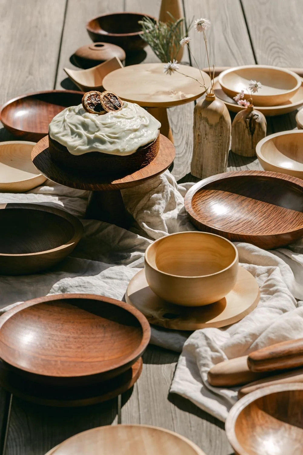 A rustic wooden table displays empty wooden bowls, a vase with dried flowers, and a cake with white frosting and dried fruit on top.