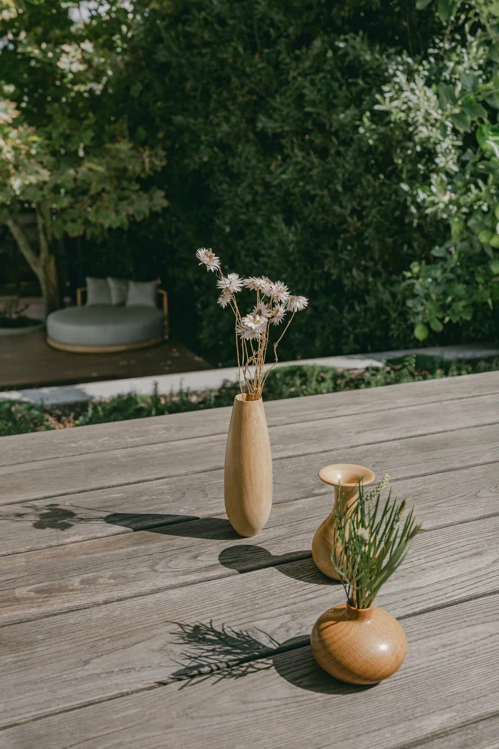 Three wooden vases with dried flowers and green foliage on wooden decking, with a garden and a white outdoor sofa in the background.