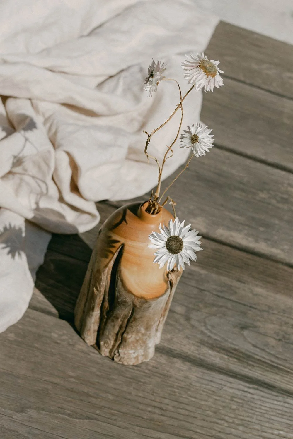 A small bouquet of white flowers in a Huon Pine Limb Wood timber vase on a wooden surface. A soft, light-colored fabric is draped in the background.