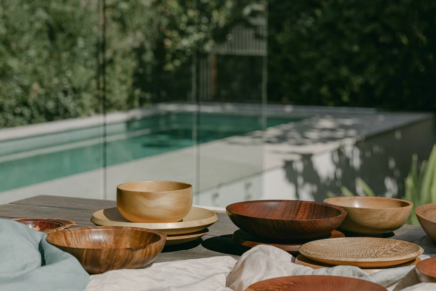 Wooden bowls and plates on a table outdoors near a swimming pool with greenery in the background.
