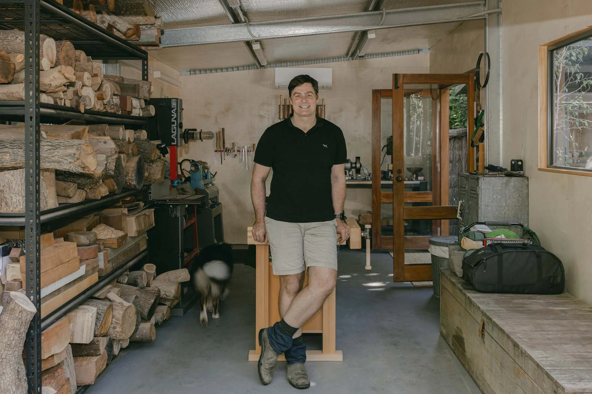 Australian wood turner, Andrew McNamara, standing in his woodworking workshop, with a black and white dog near his feet. The shop has shelves filled with chopped wood on the left, tools and equipment on the back wall, and a window on the right.