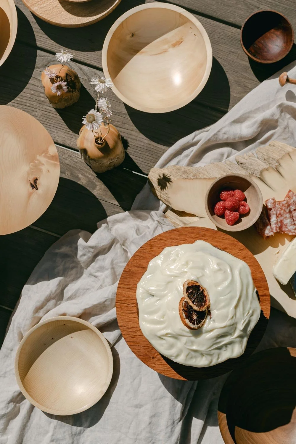 A rustic outdoor table setup with wooden bowls, a creamy cheese dish garnished with dried citrus slices, fresh raspberries, and a variety of decorative elements like flowers and cloth.