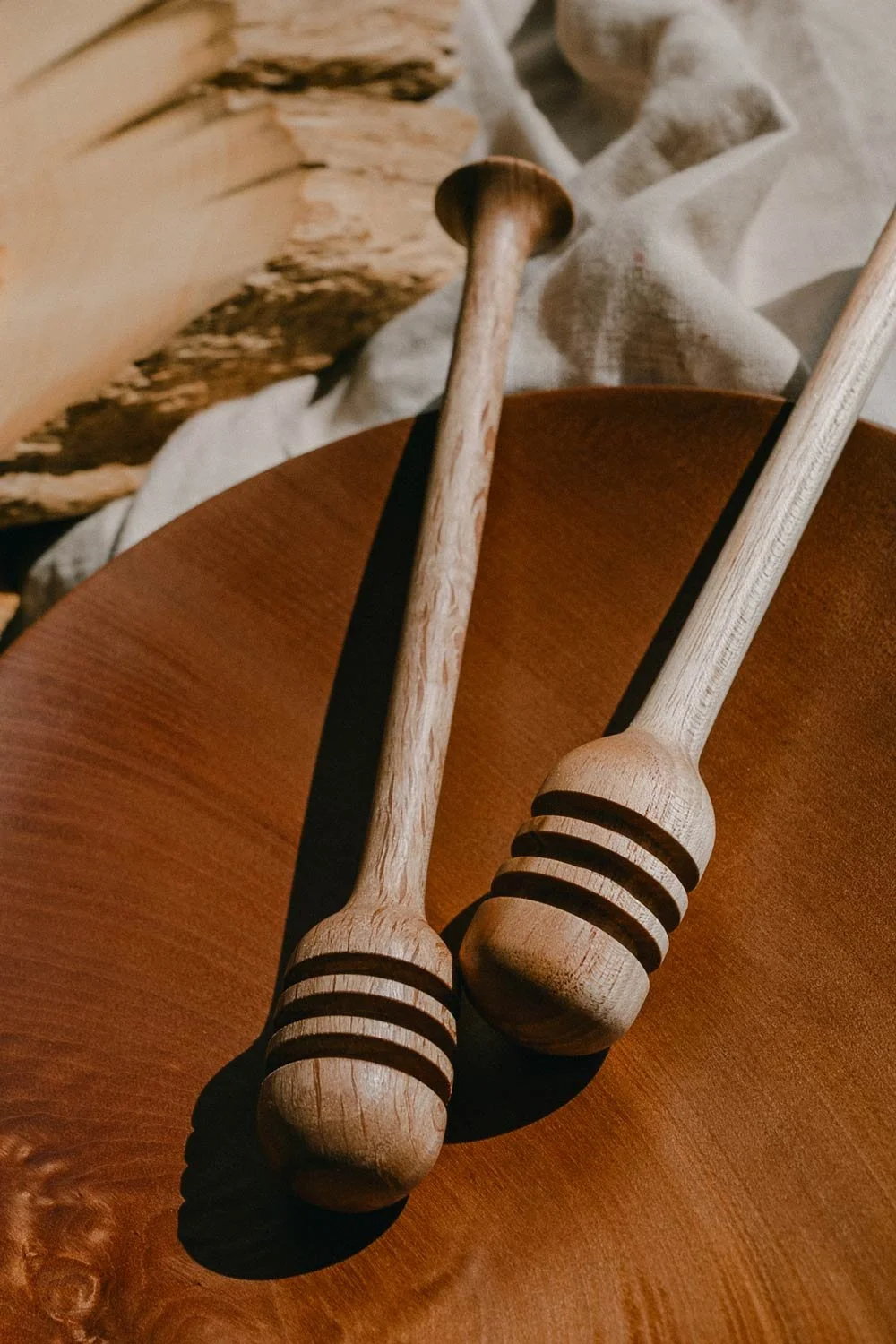 Two wooden honey dippers resting on a round wooden surface, with a piece of driftwood and cloth in the background.