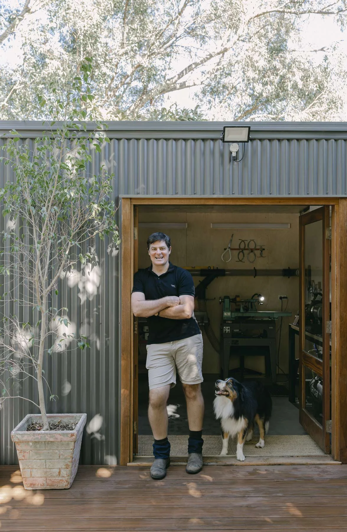 Andrew McNamara, Perth wood turner, in his wood workshop doorway with his dog beside him, inside the workshop tools are visible. A potted tree is outside.