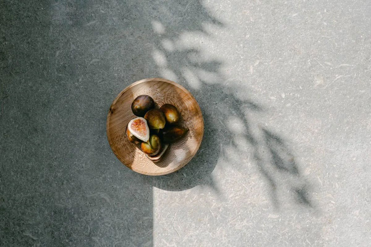 A wooden bowl containing ripe figs on a textured gray surface with shadows of leaves cast on the surface.