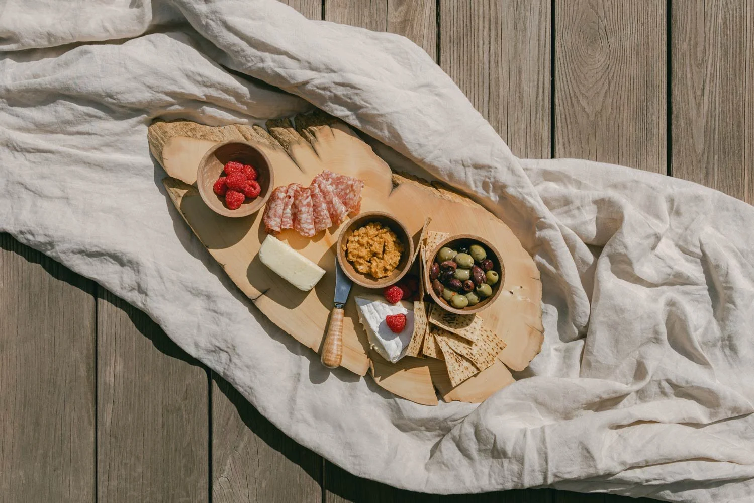 Timber platter/board with charcuterie of cheese, meats, olives, raspberries, crackers, and spread on a wooden surface, partly covered with a white cloth.