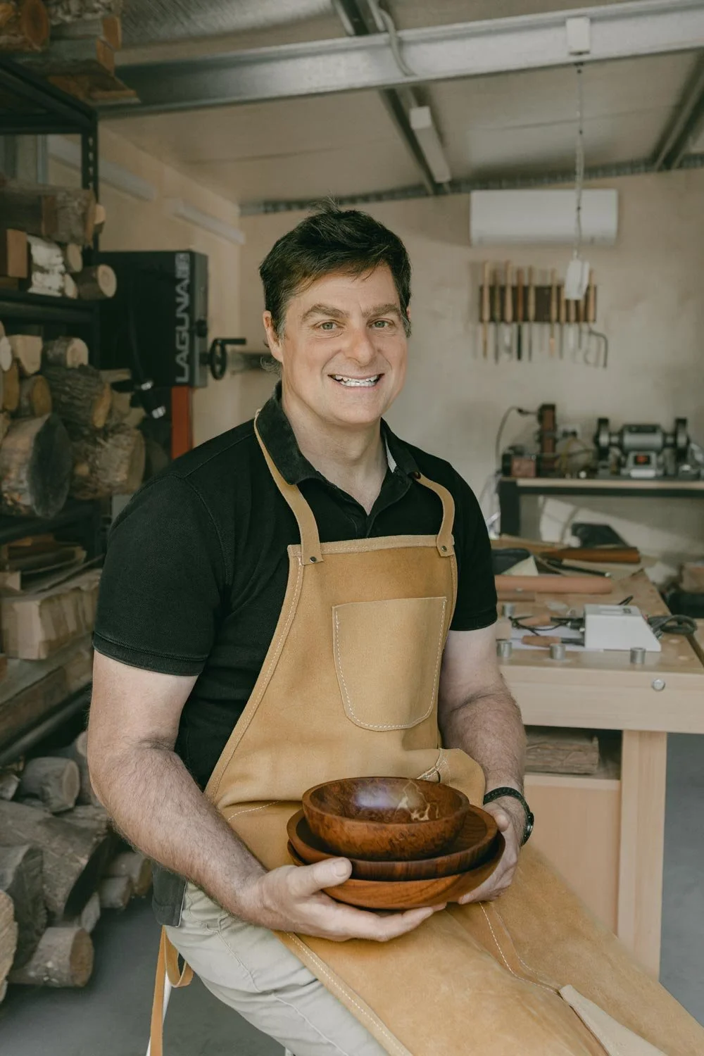 Andrew McNamara, Perth wood turner, seated on a stool, smiling, in a black shirt and beige apron holding wooden bowls in his woodworking workshop.